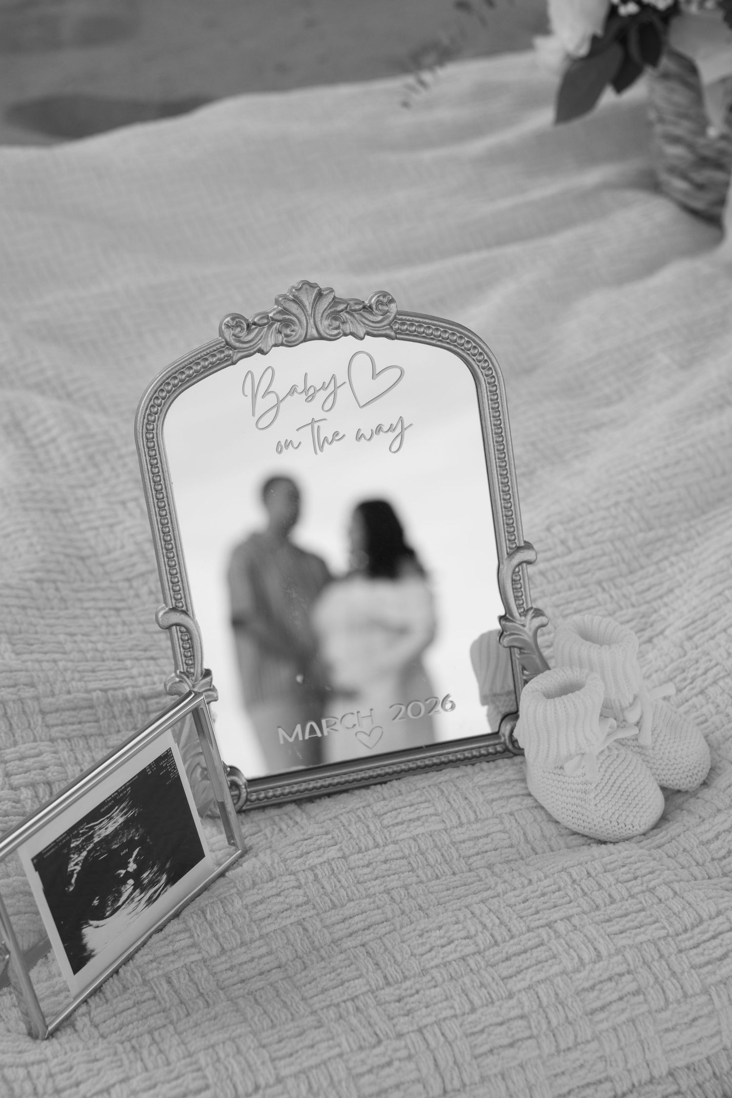 Black and white photo of a decorative mirror with baby announcement, ultrasound photo, and tiny knit booties on a bed.