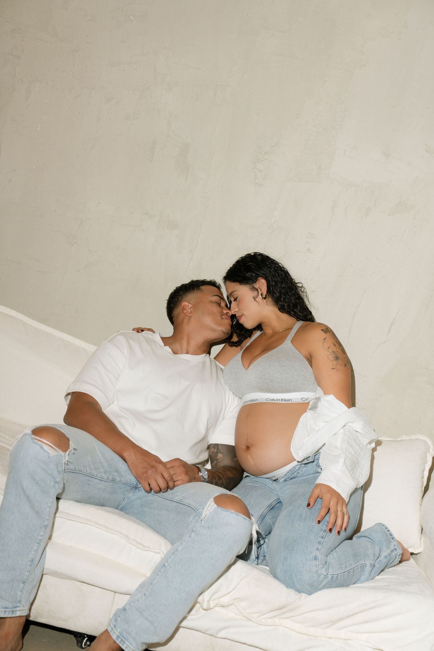 Expecting couple sharing a kiss on white couch, woman's baby bump visible, wearing matching jeans outfits.