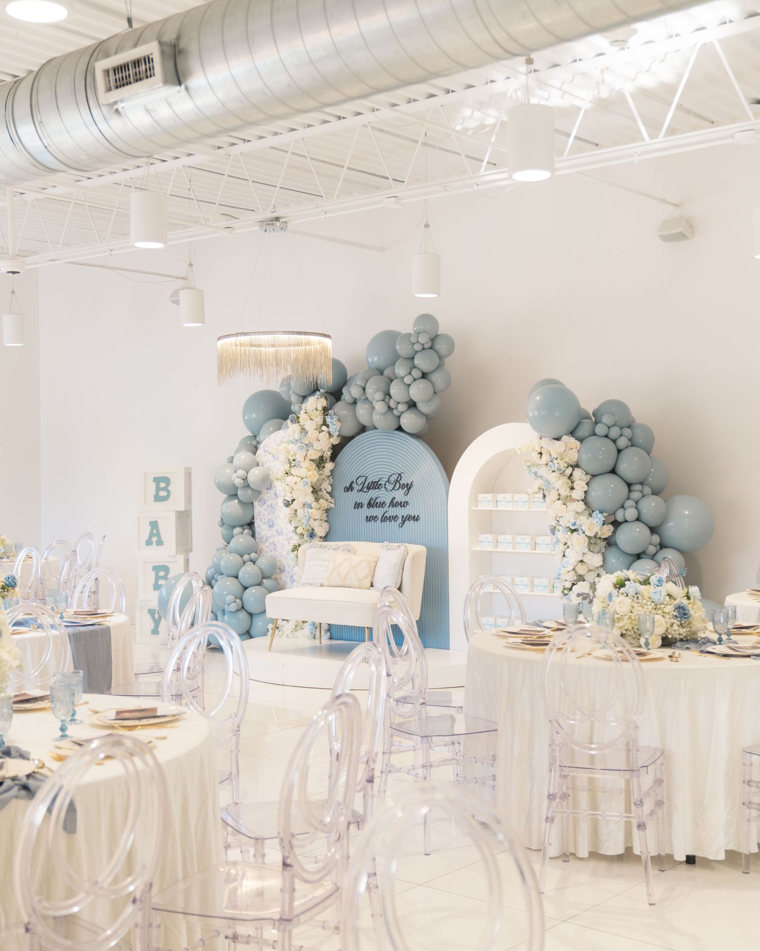 Elegant wedding reception venue with white and blue balloon decor, ghost chairs, and modern industrial ceiling details.