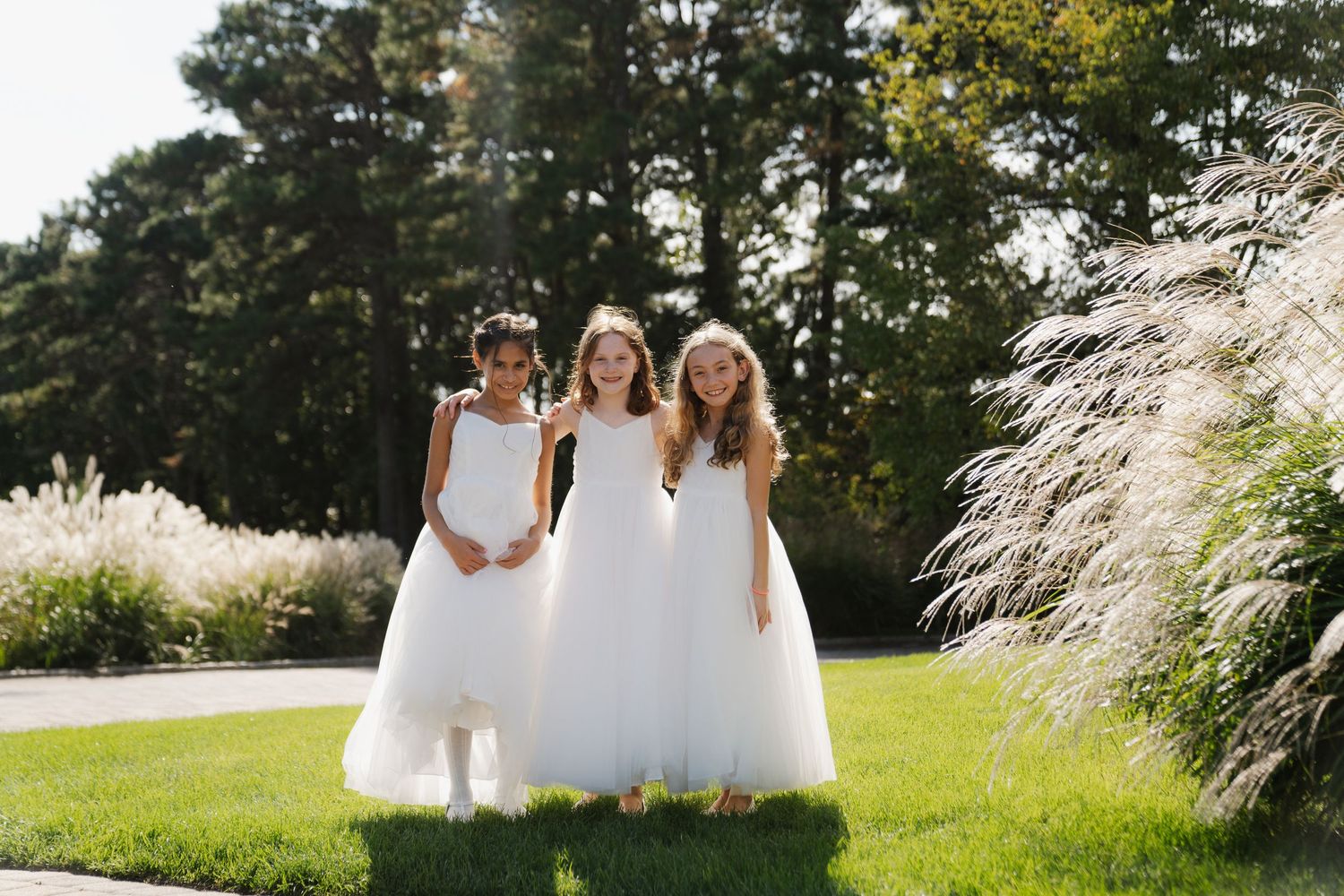 A group in white dresses walks through a sunny garden setting with trees and grass in the background.