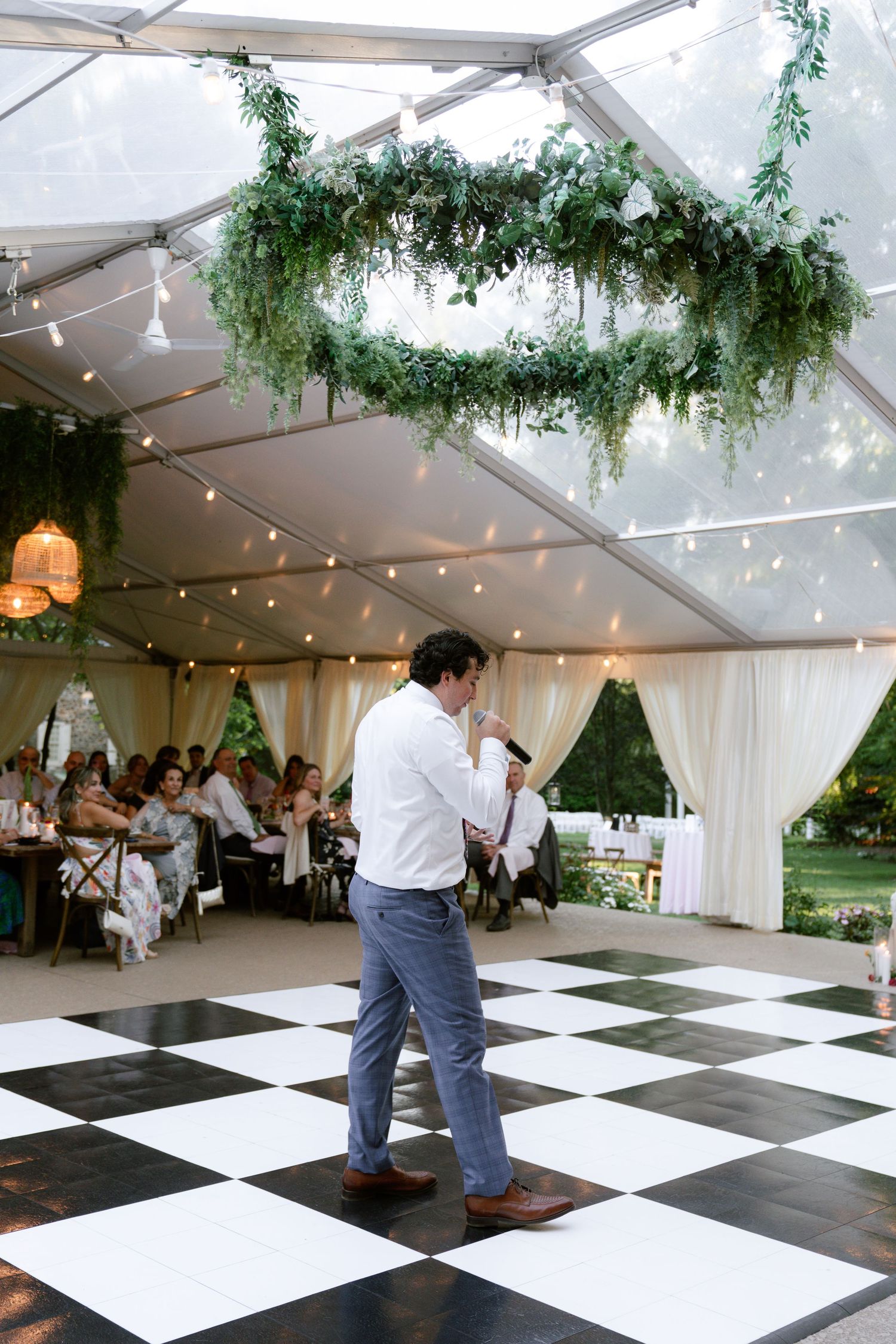 A wedding reception tent with black and white checkered dance floor and hanging greenery chandelier.