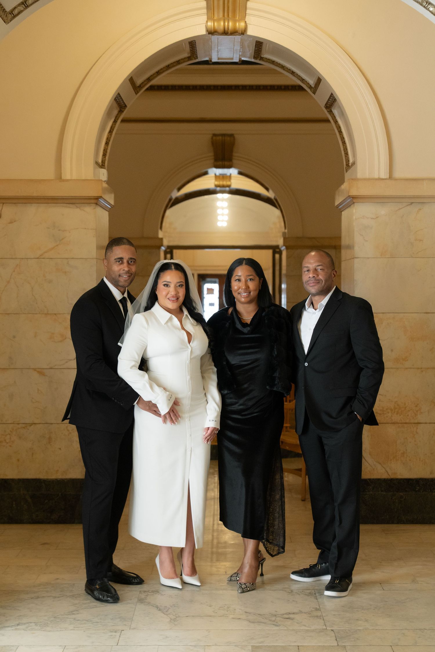 Four people in formal attire pose together under elegant arched hallway in classic architectural setting.