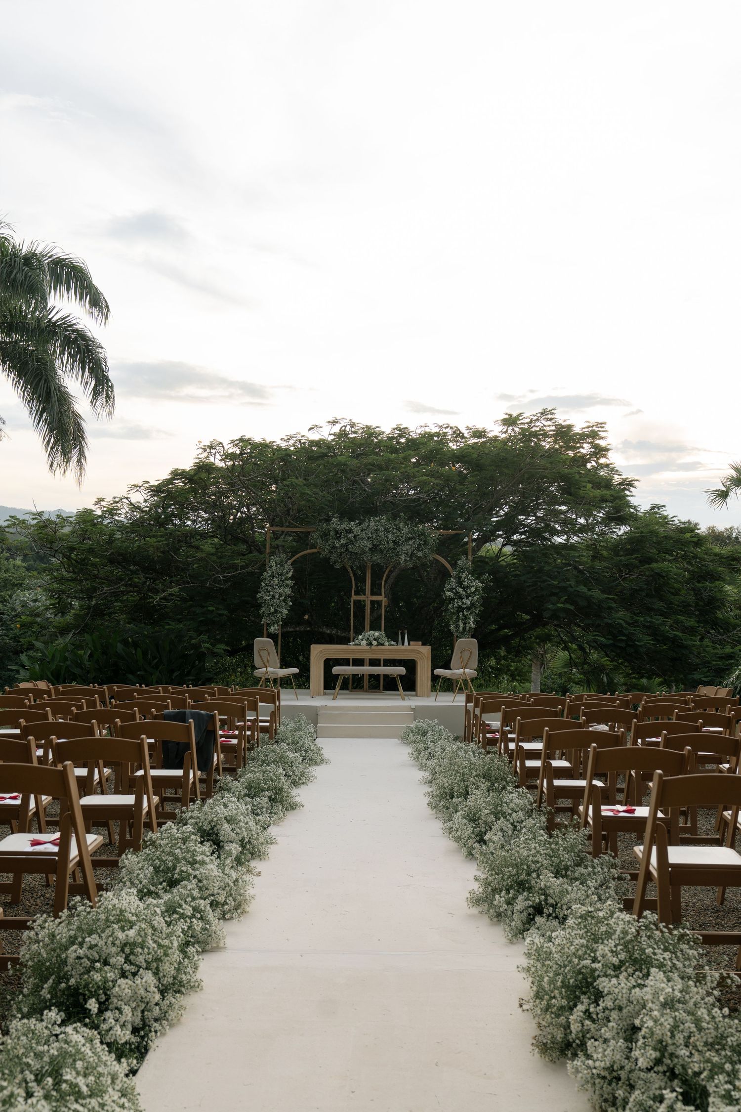 An outdoor wedding ceremony setup with wooden chairs and floral aisle decorations under palm trees.