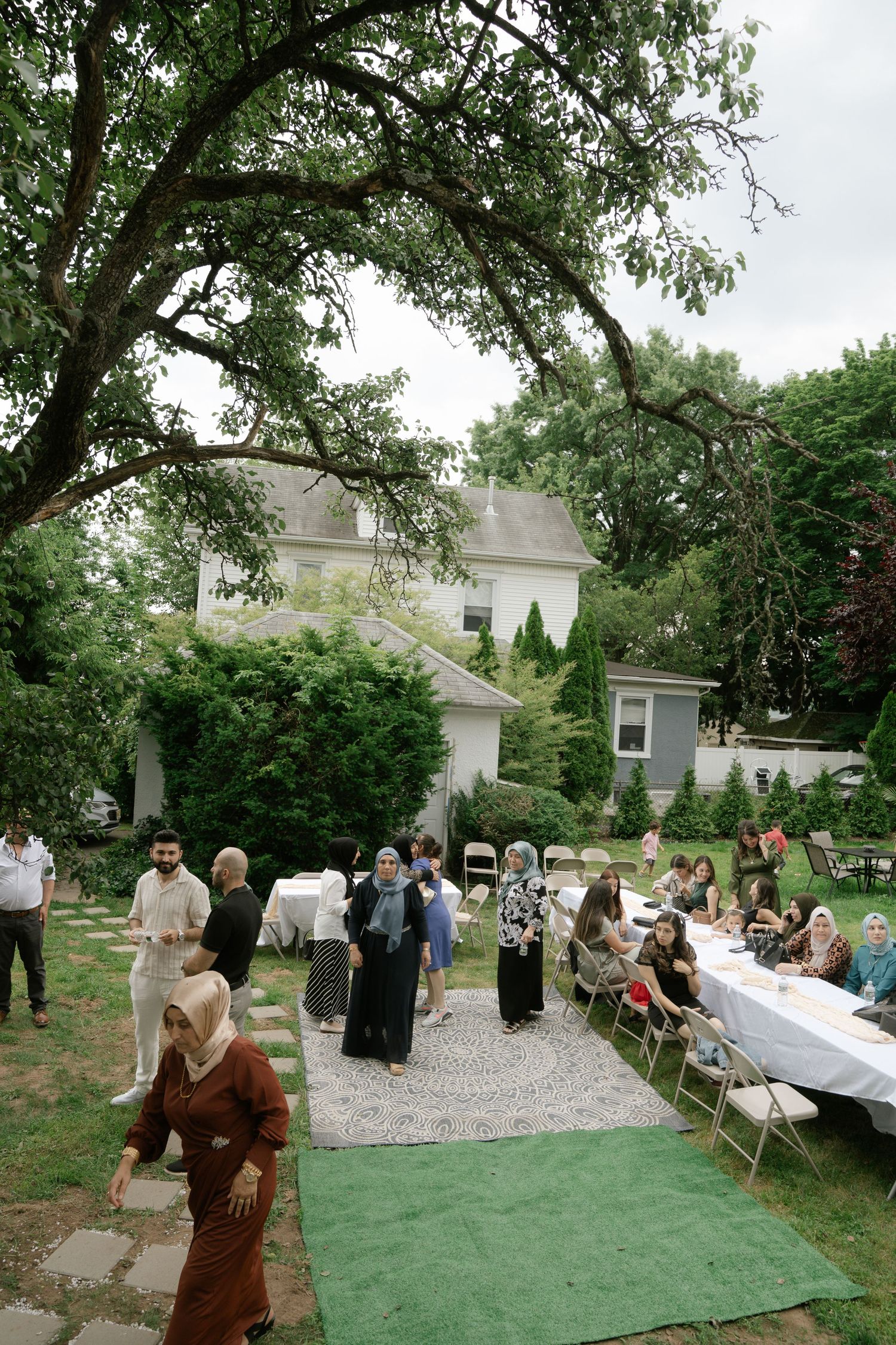 Outdoor garden reception with guests mingling on a green carpet under large tree branches.