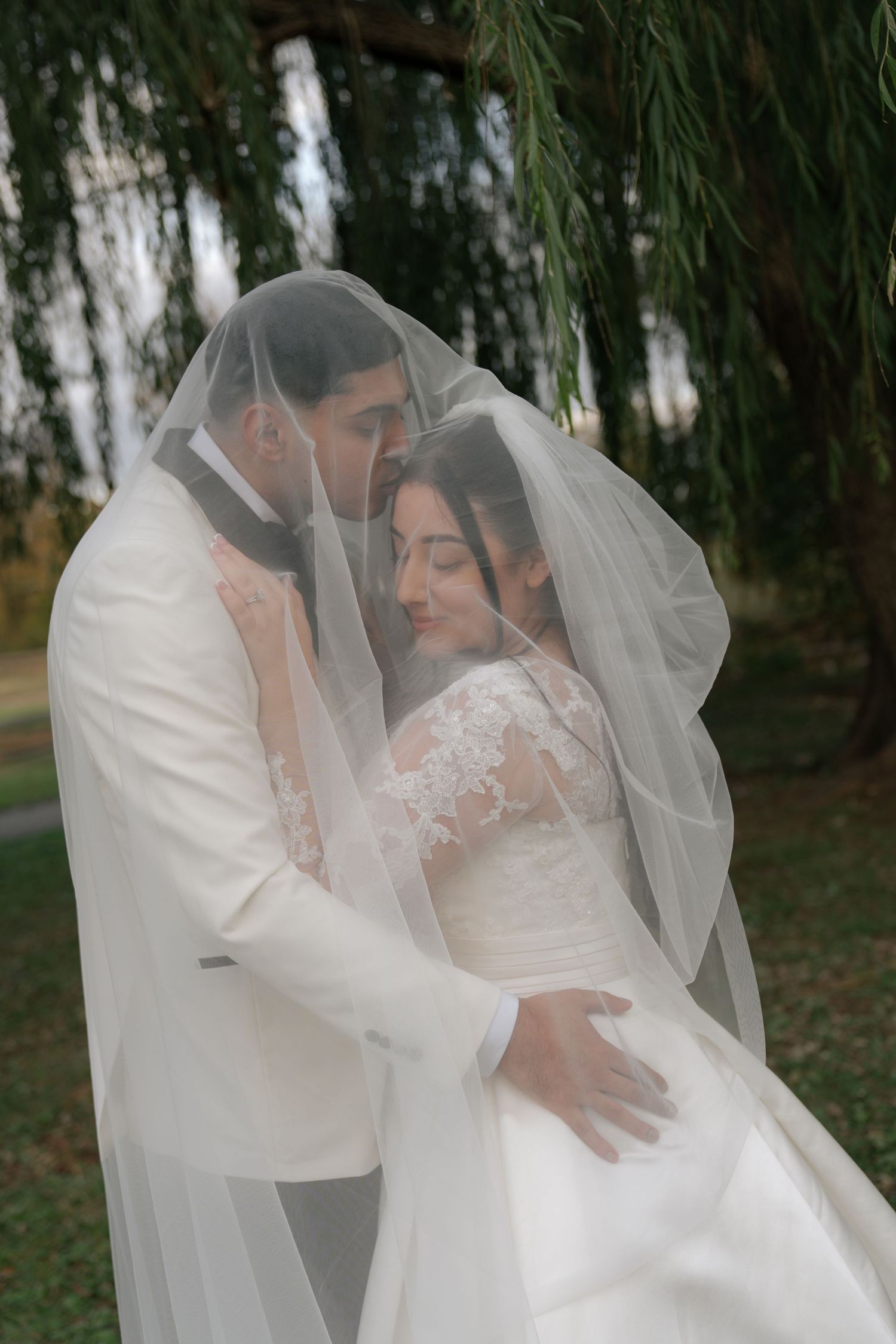 Romantic wedding portrait under a veil with willow trees in the background.