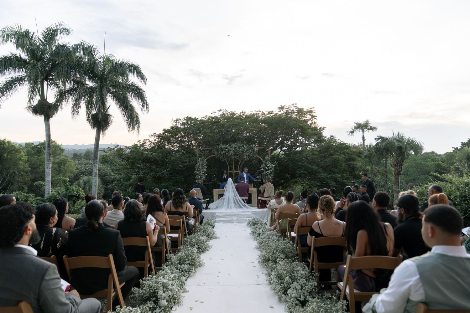 Outdoor wedding ceremony setup with white chairs and aisle runner framed by palm trees.