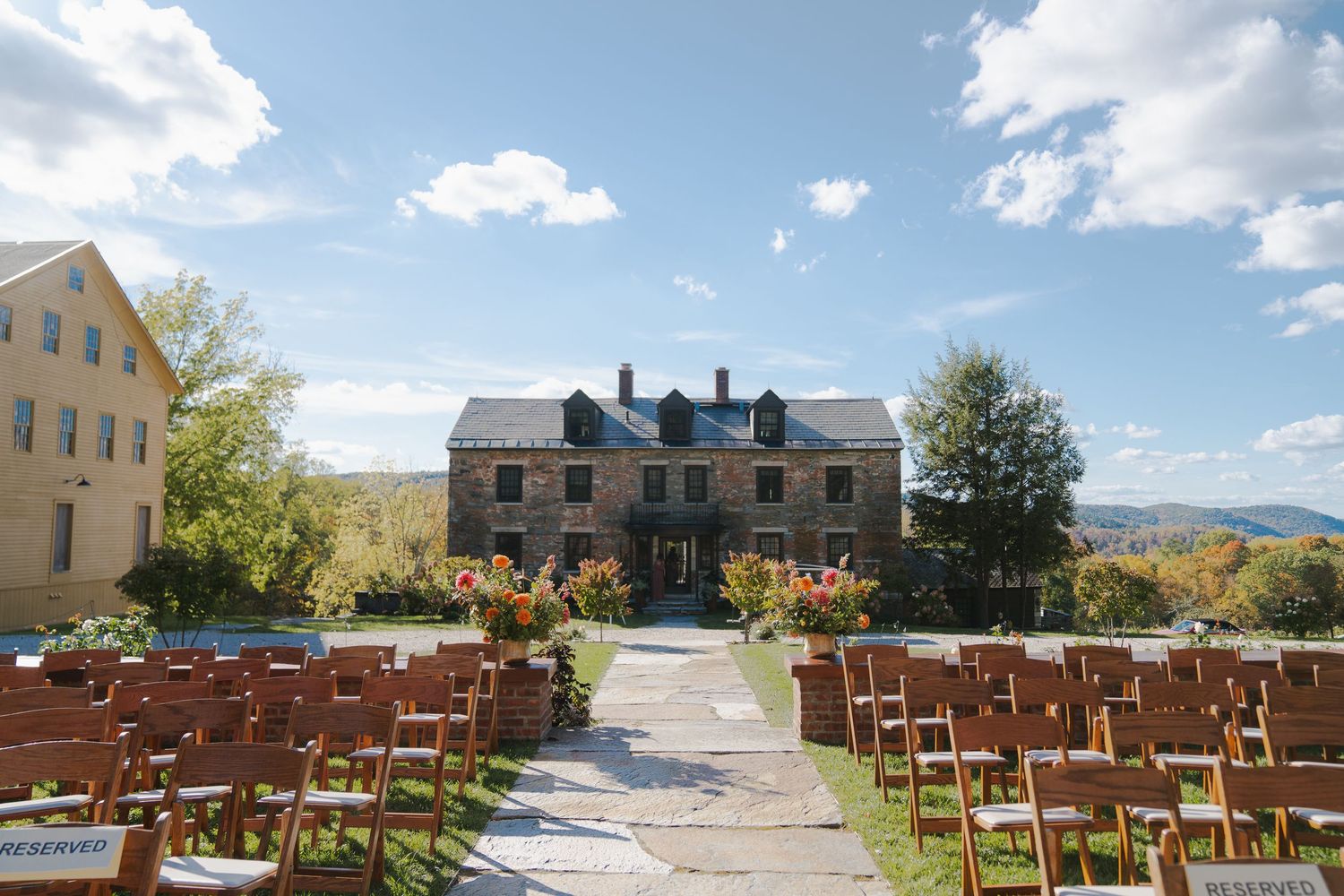 Outdoor wedding ceremony setup with wooden chairs facing historic buildings under blue sky.