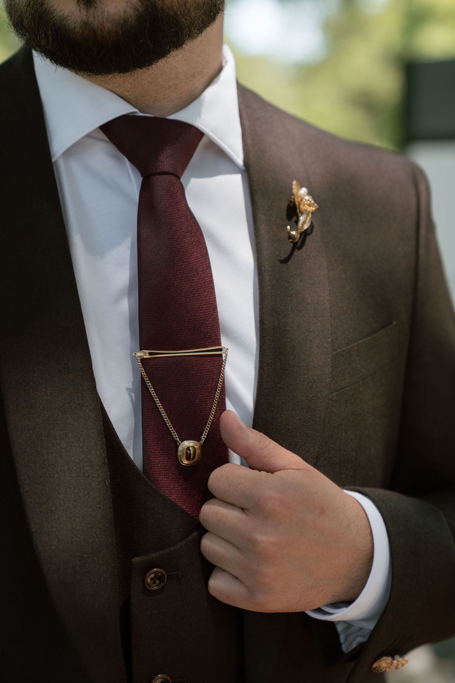 Groom in brown suit with burgundy tie, gold tie bar, chain pendant, and floral boutonniere at wedding.
