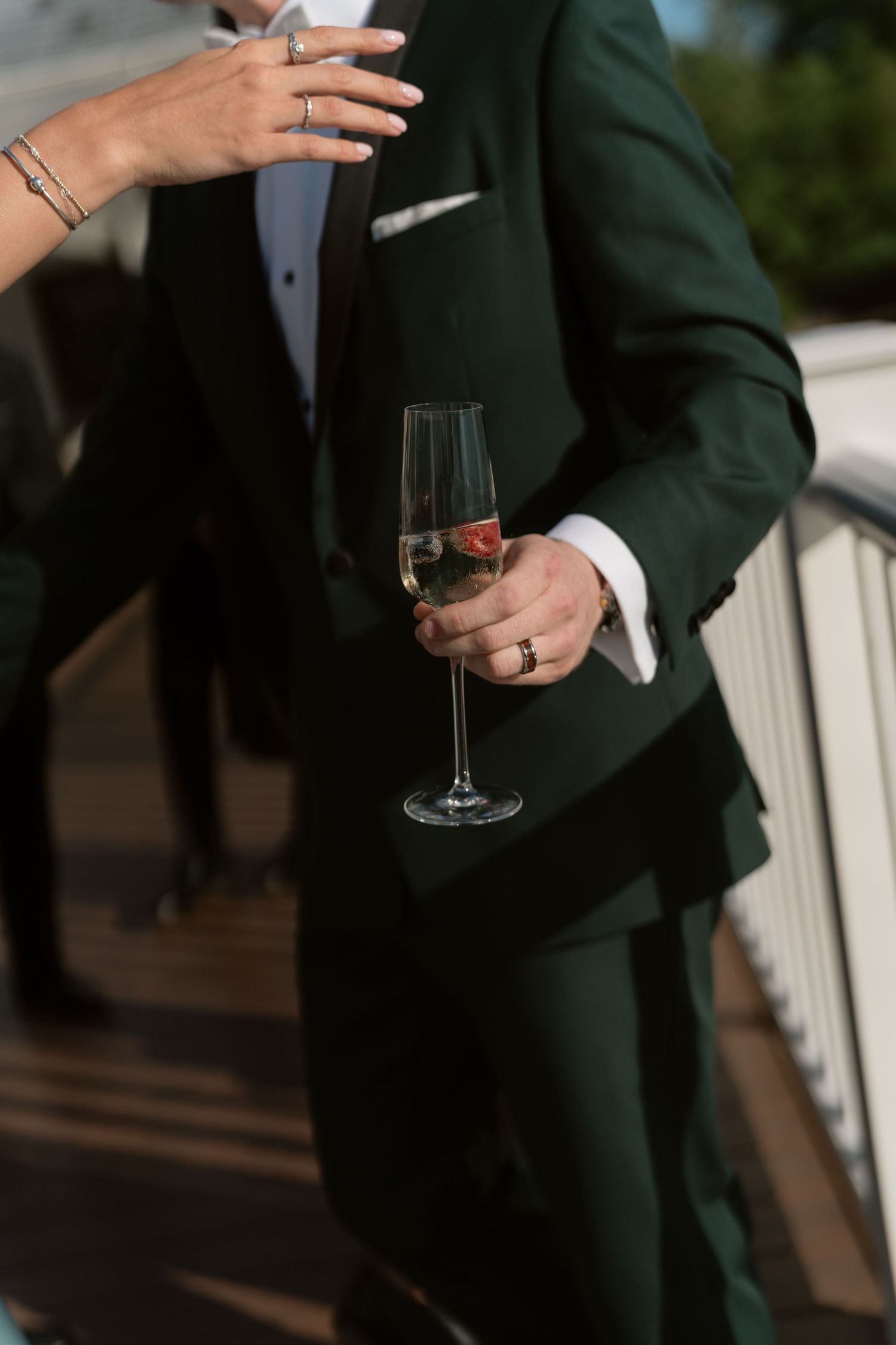 Groom in dark green suit holds champagne flute with strawberry while guest places hand on his shoulder.