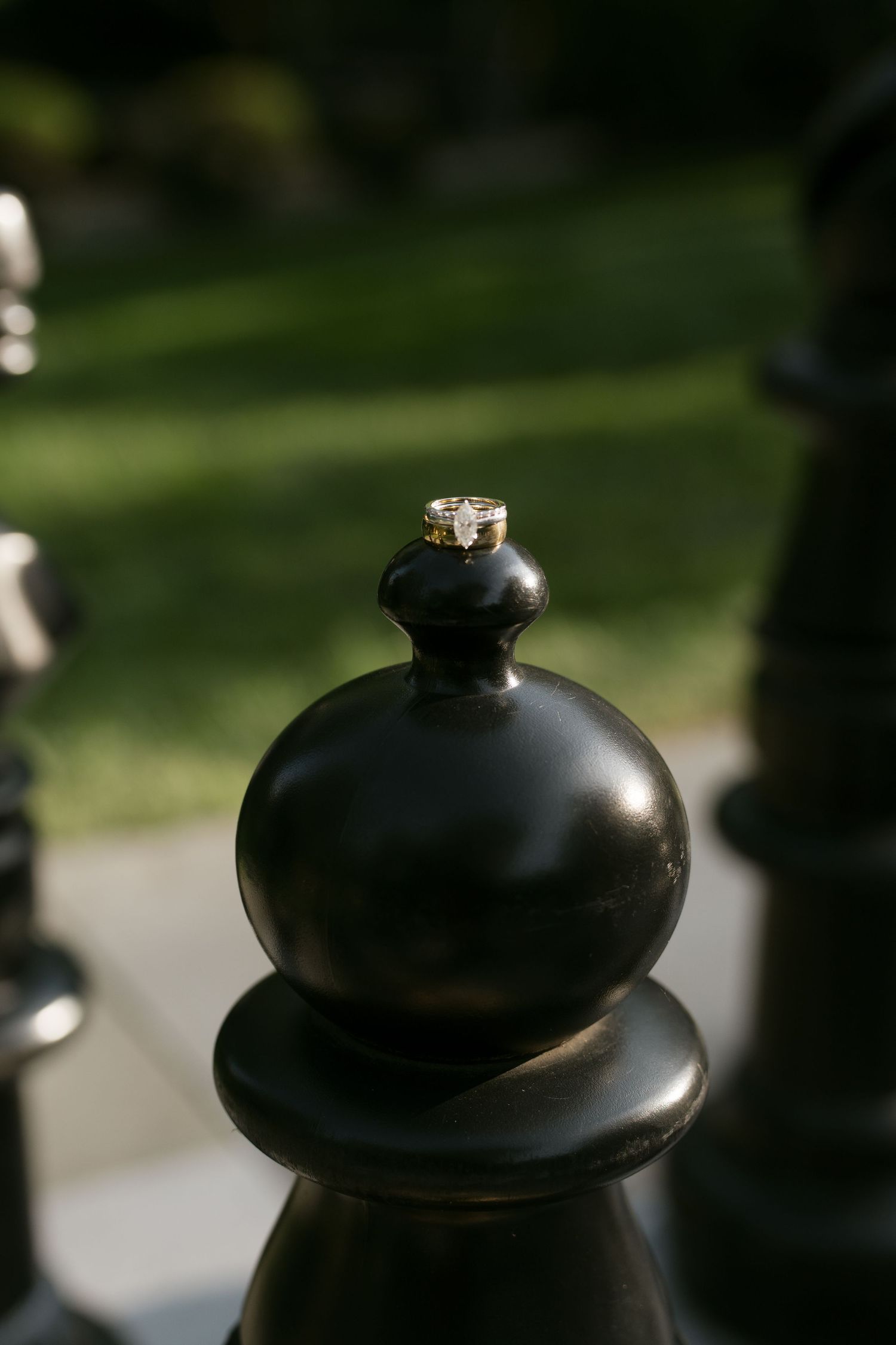 A gold wedding ring resting atop a large black chess pawn piece outdoors with green grass in the background.