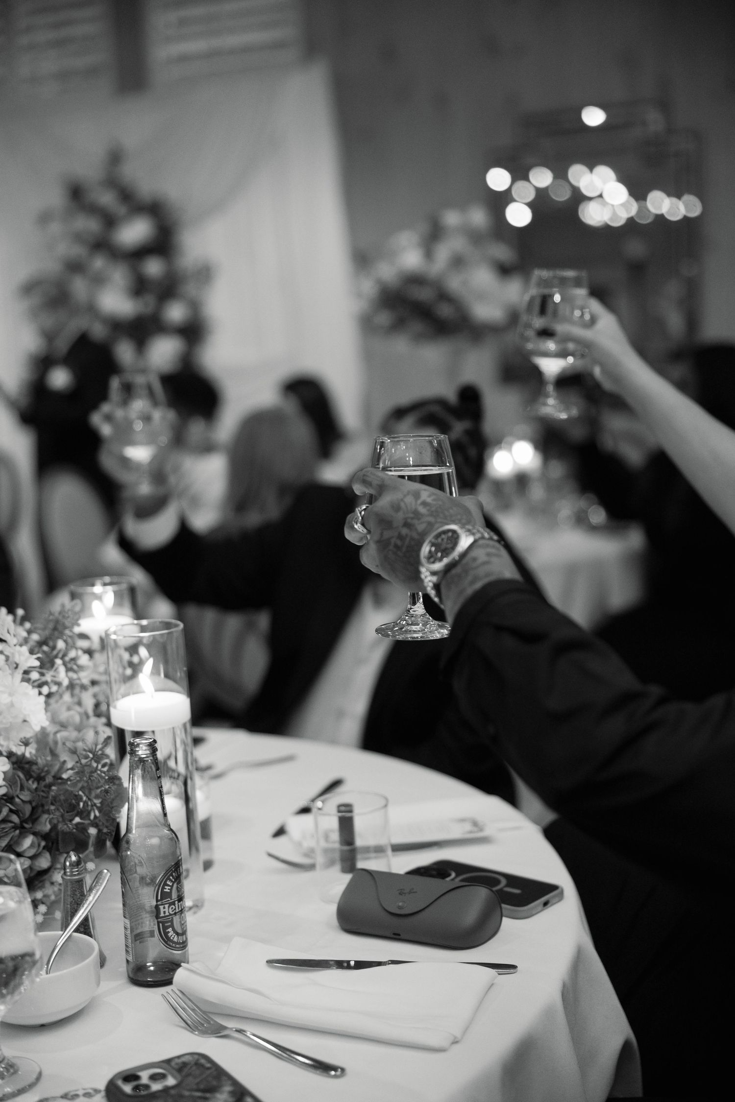 Black and white photo of wedding guests raising champagne glasses in toast at candlelit reception table.