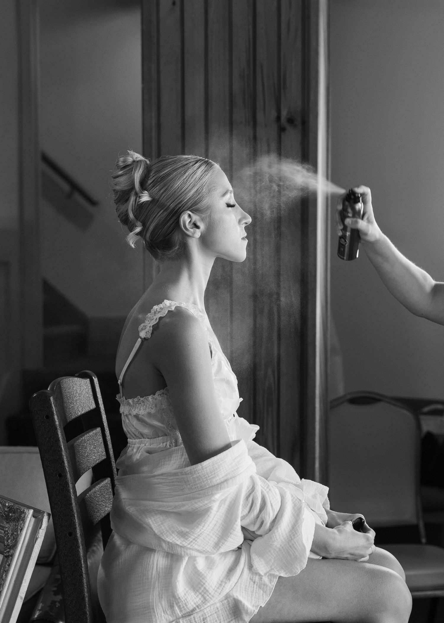 Black and white photo of a bride getting hairspray applied while seated in a chair, eyes closed, in a white dress.