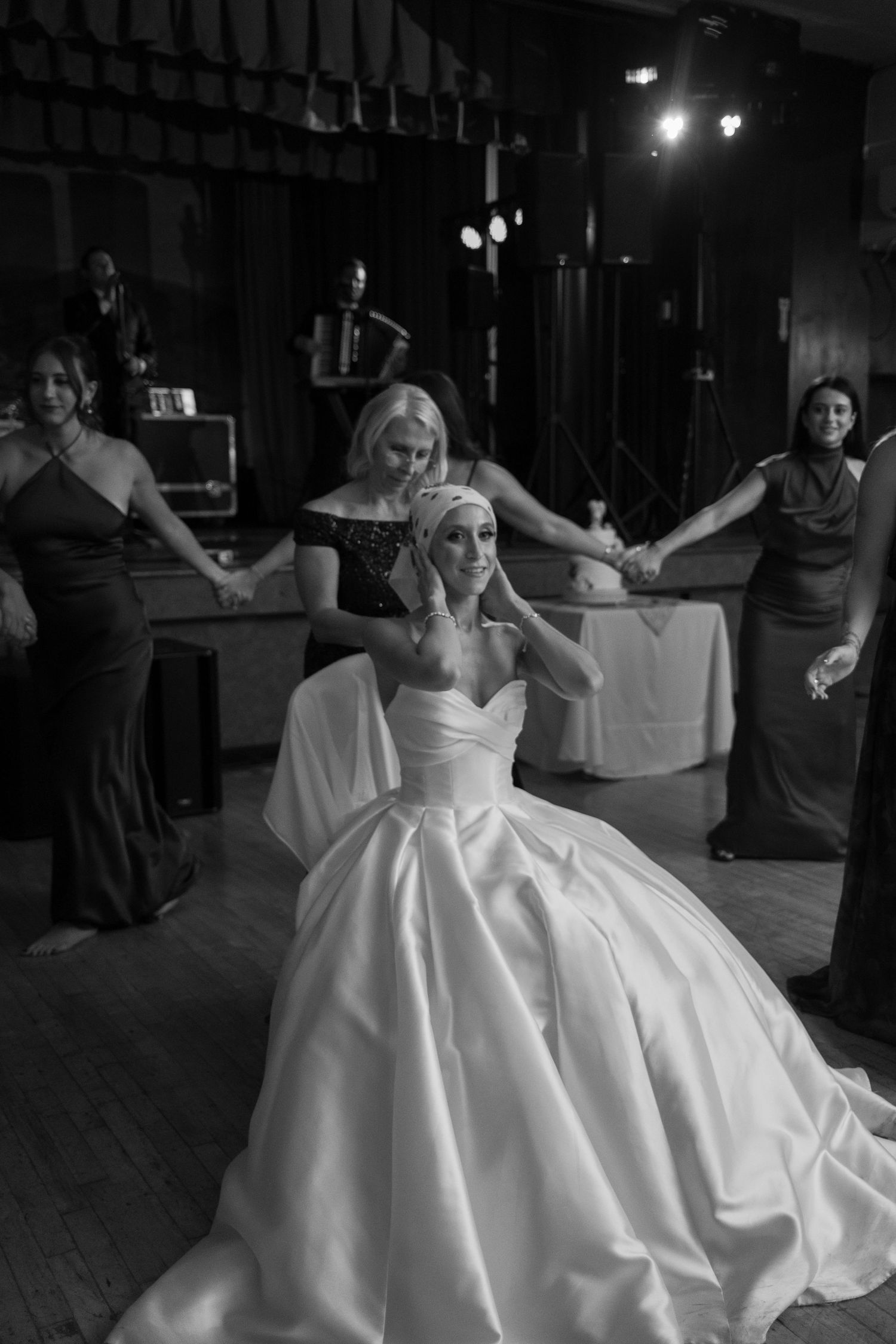 Bride in a ballgown sits on the dance floor surrounded by bridesmaids holding hands in black and white photo.