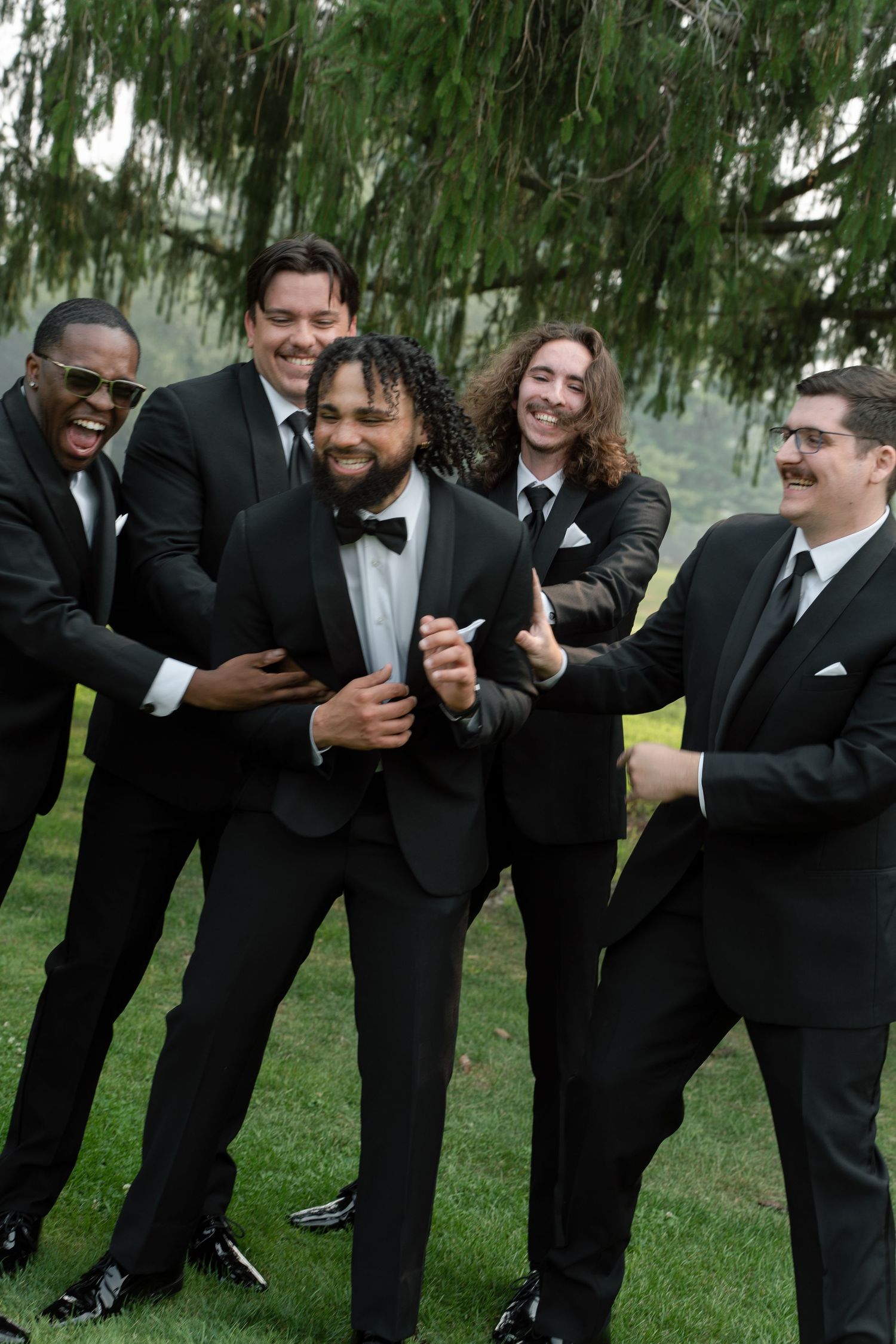 Groom in black tuxedo laughs joyfully surrounded by groomsmen in matching suits outdoors.