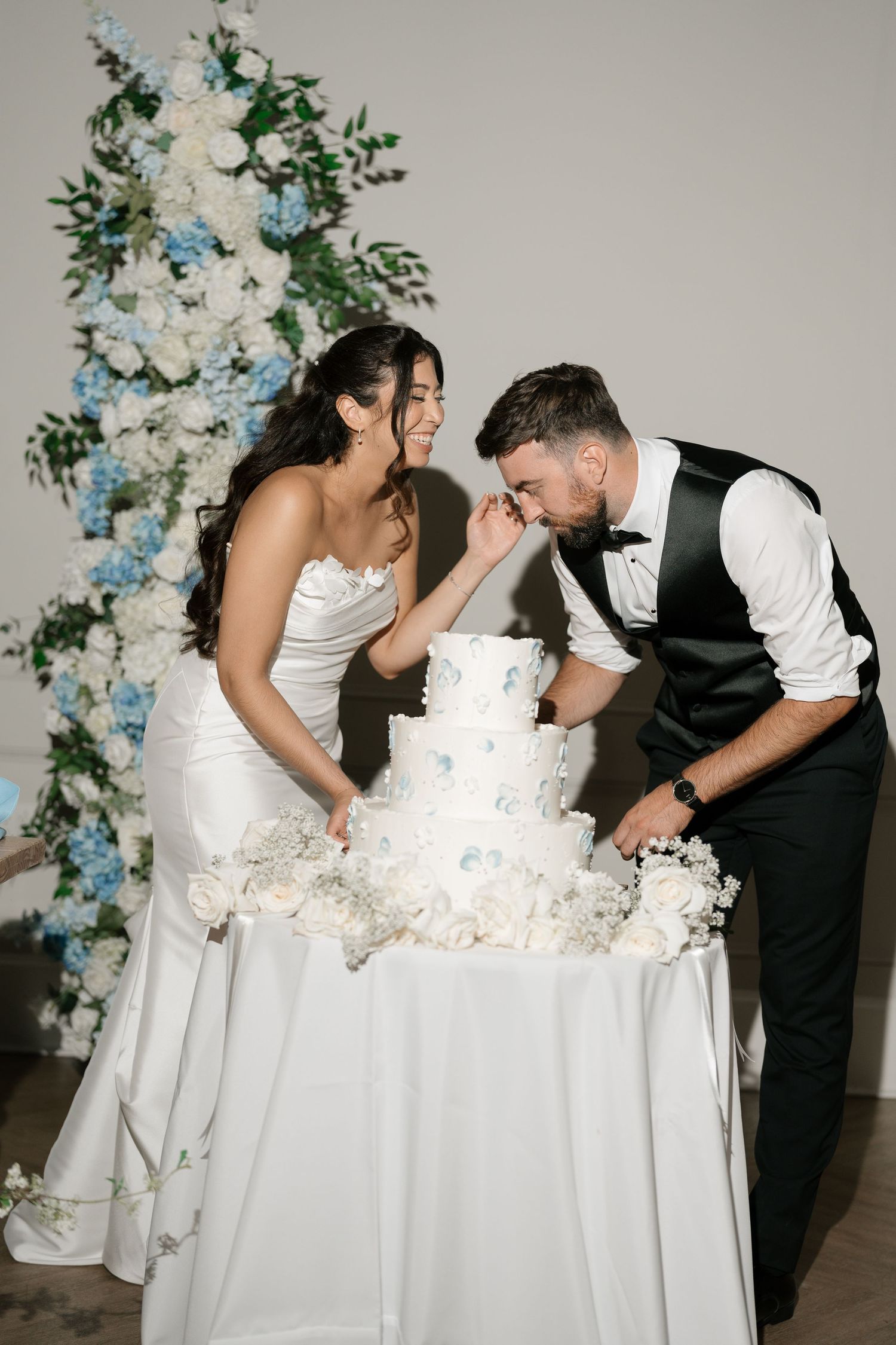 Bride and groom cutting three-tier white wedding cake with blue and white floral backdrop.