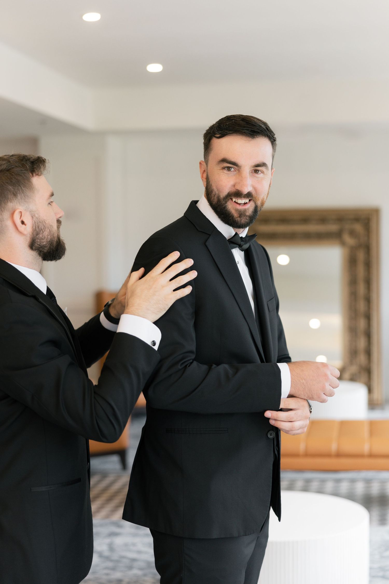 Groomsman helping groom in black tuxedo and bow tie prepare for wedding ceremony.