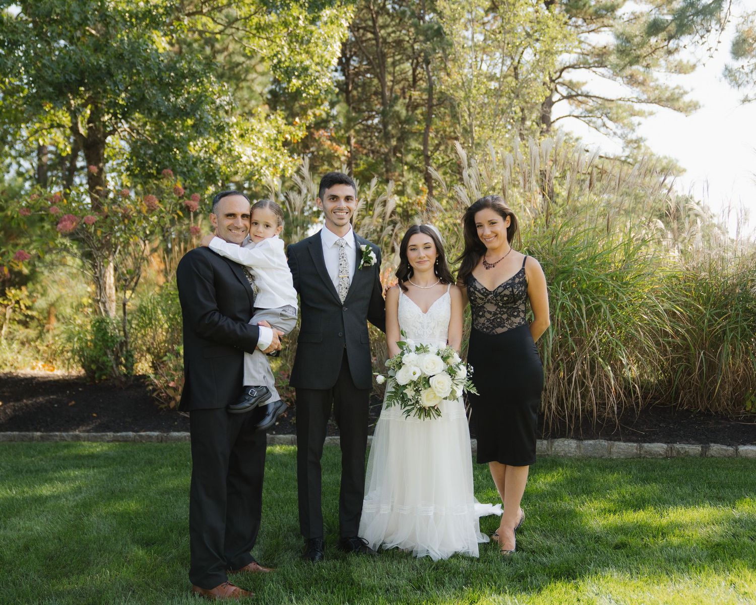 Wedding party poses together outdoors in formal attire with white bouquets against a natural garden backdrop.