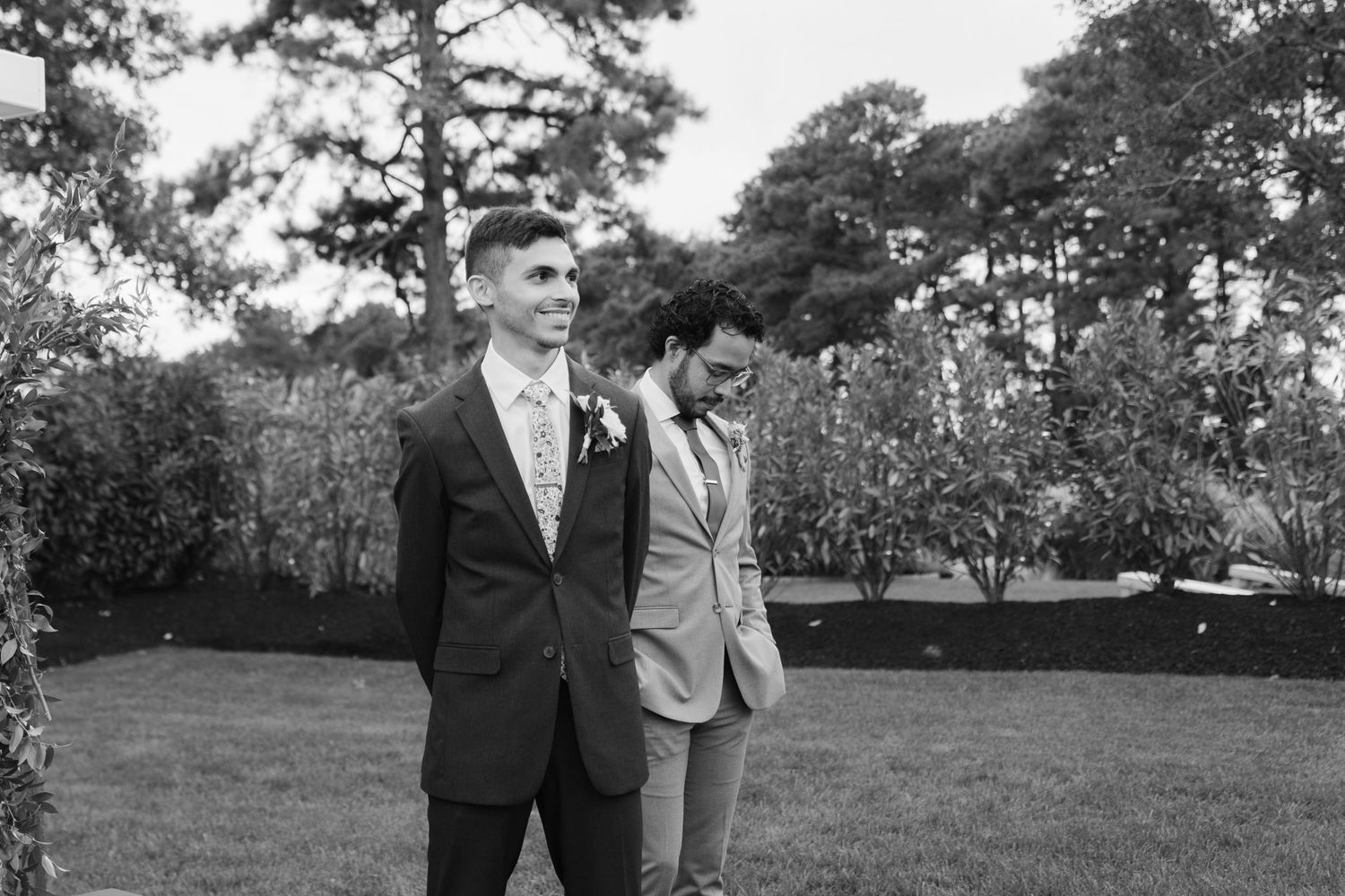 Two people in formal suits stand together on a lawn with tall pine trees in the background.