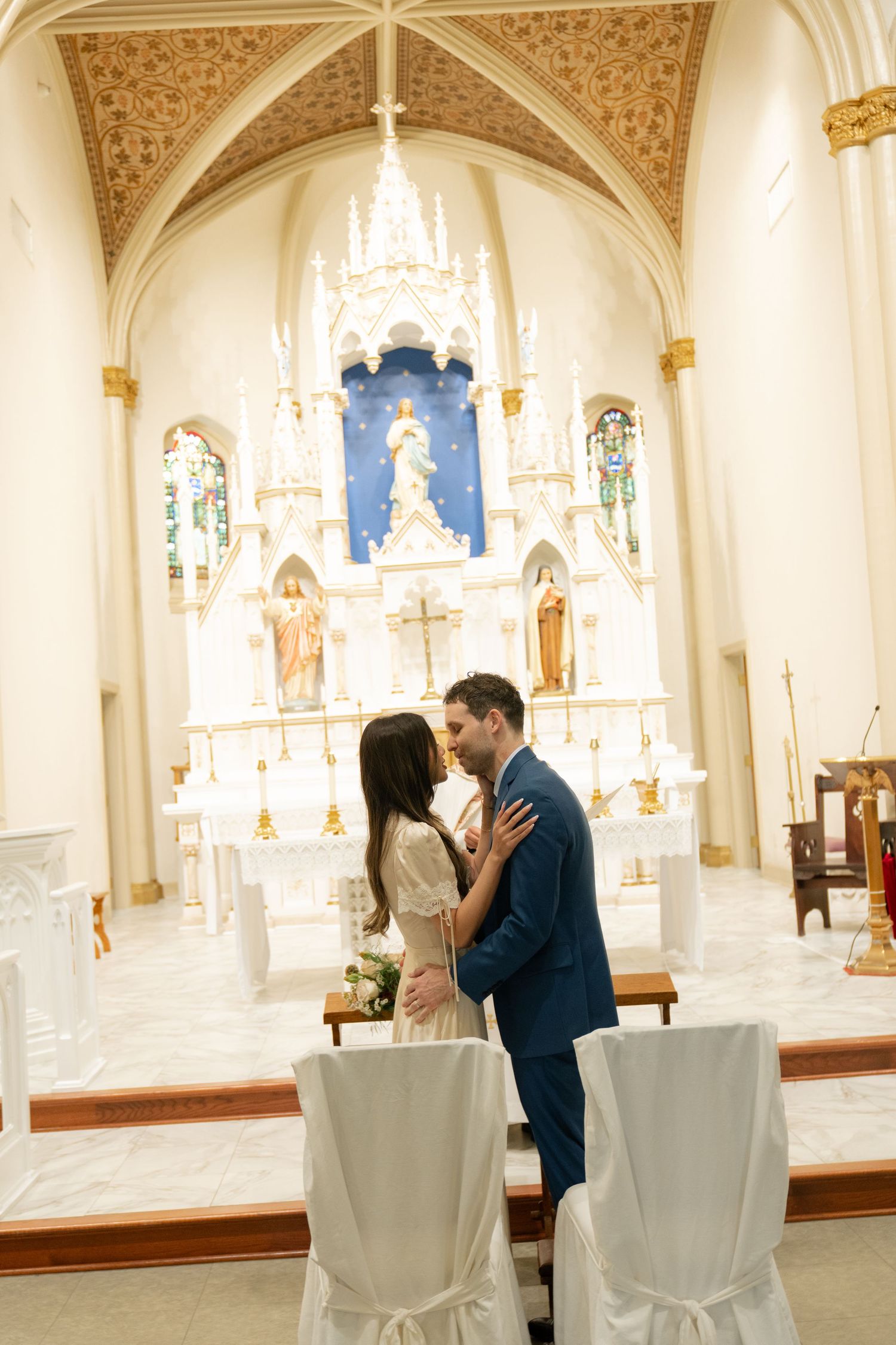 A couple embraces near the altar of a traditional Catholic church with ornate decor.