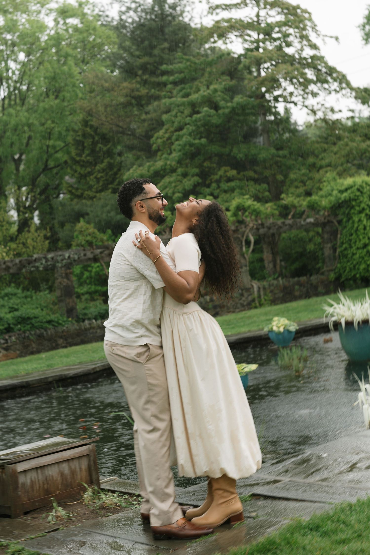 A couple embraces and laughs together in a lush garden near a pond, dressed in elegant cream outfits.