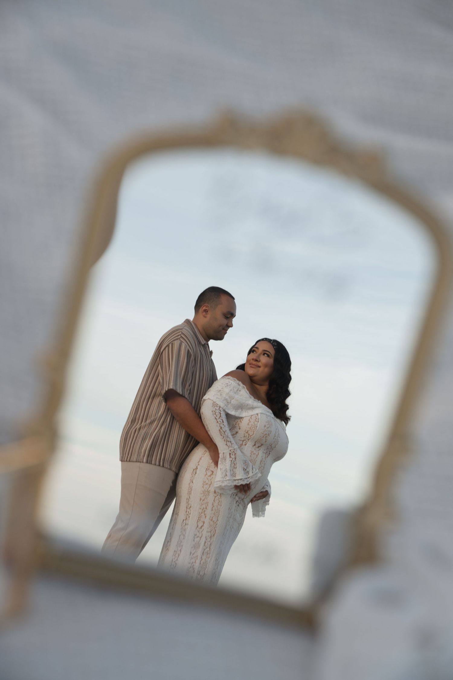 A couple embraces romantically, framed through a decorative mirror outdoors at dusk, woman in white lace dress.
