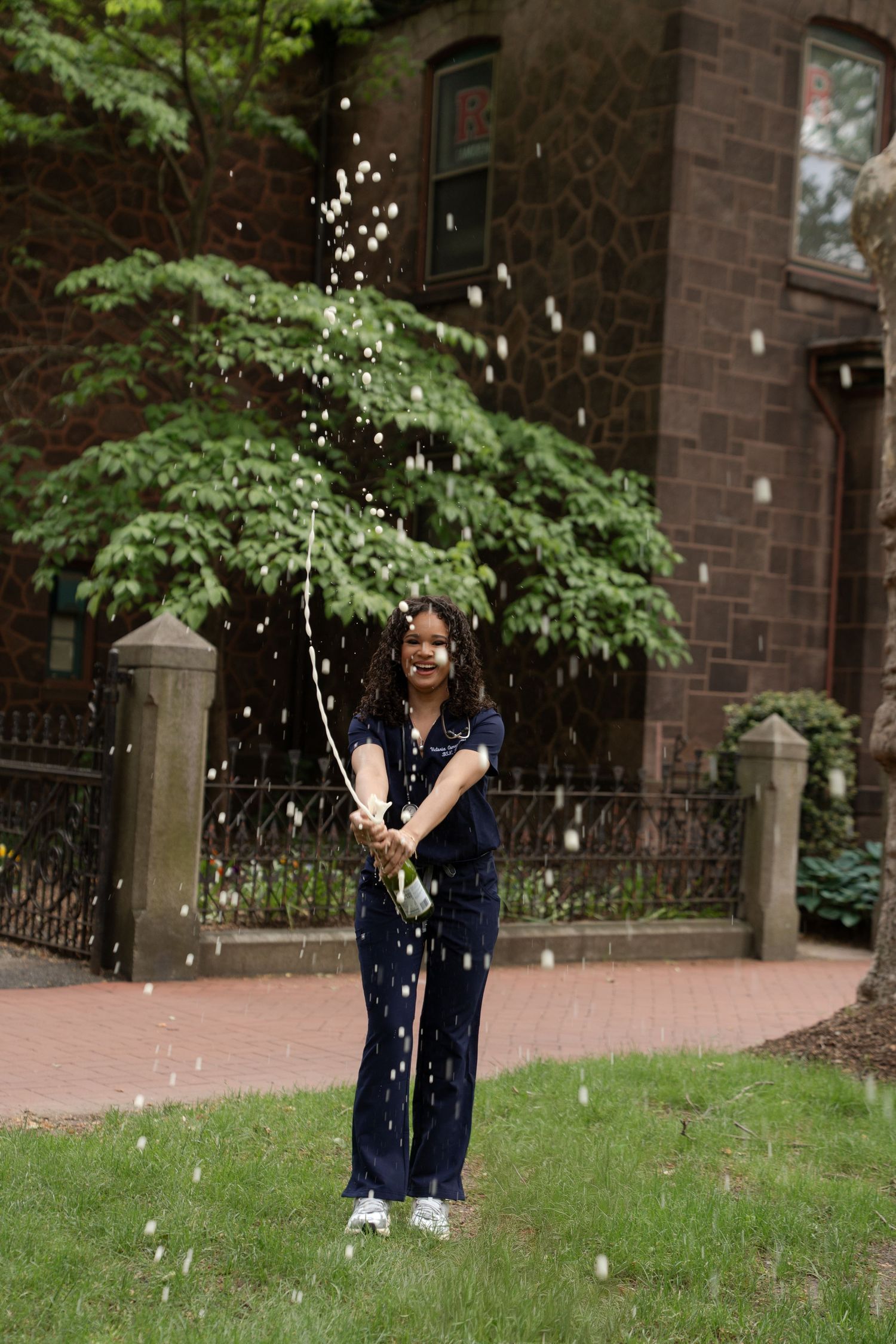 Young woman in navy scrubs celebrating by popping a champagne bottle on a green lawn outdoors.