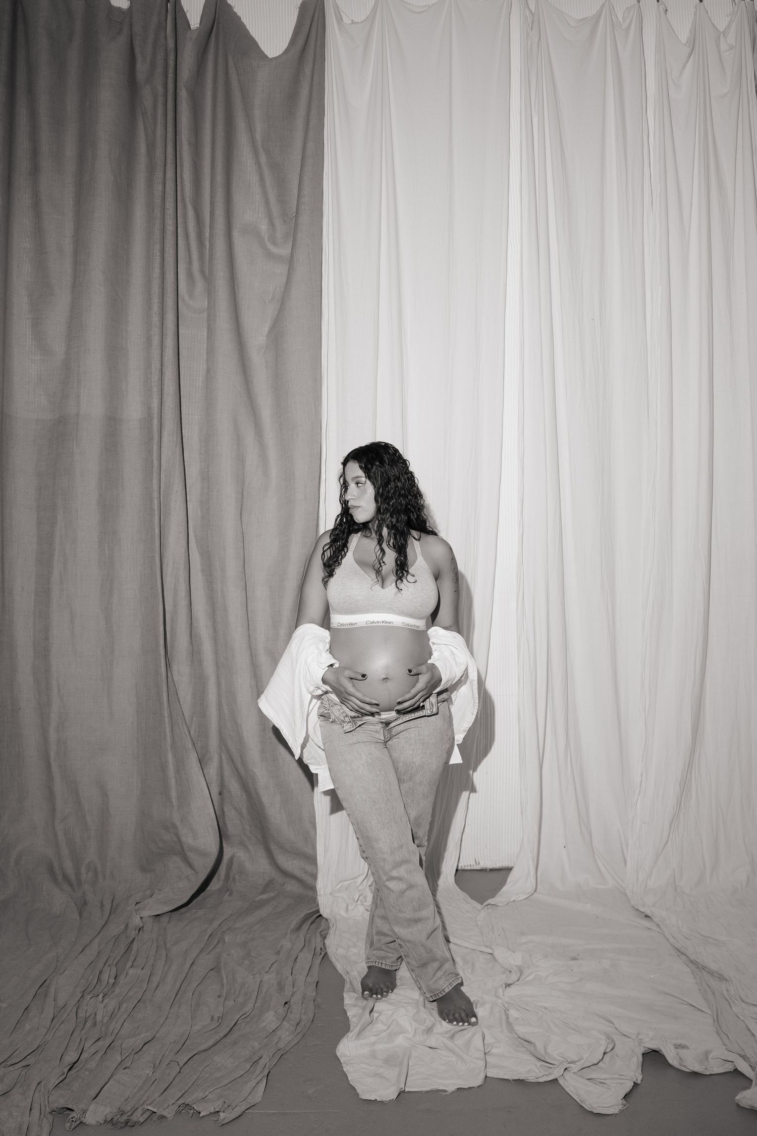 Black and white photo of a pregnant woman in a crop top and jeans, posing in front of draped fabric backdrops.