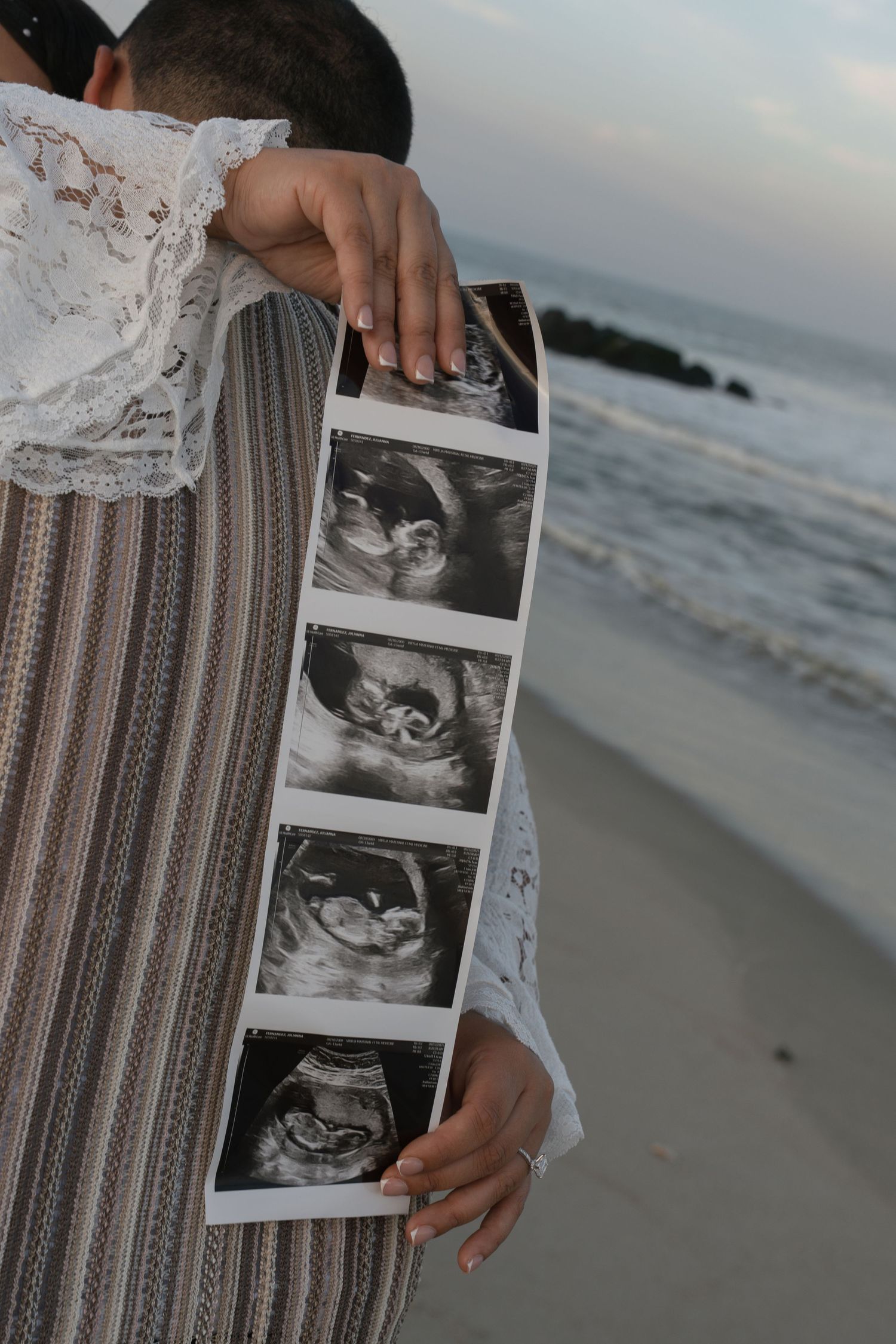 Couple embracing on beach holding ultrasound photos, pregnancy announcement at ocean shore at dusk.