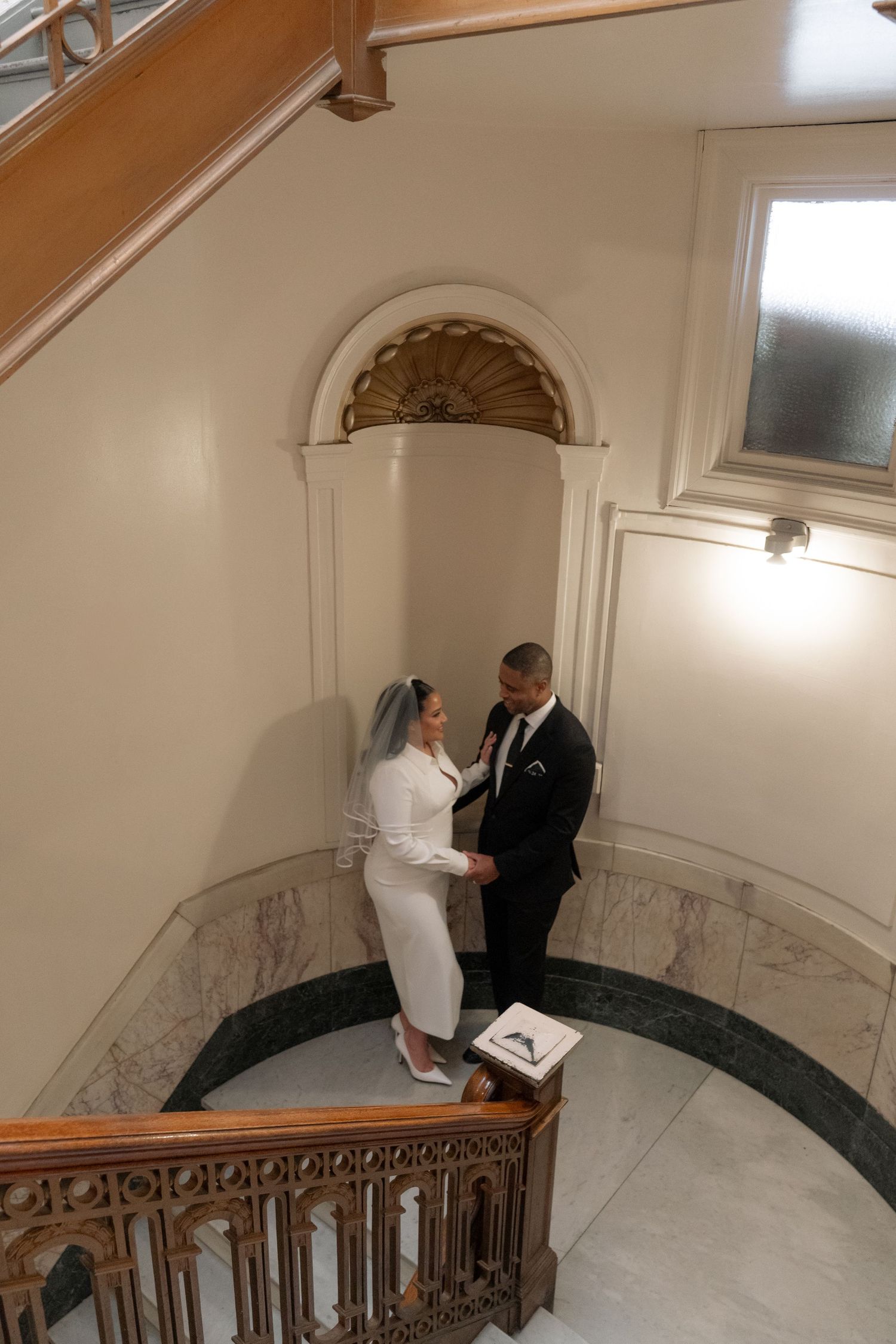A couple poses on a grand marble staircase with decorative railings and arched windows.