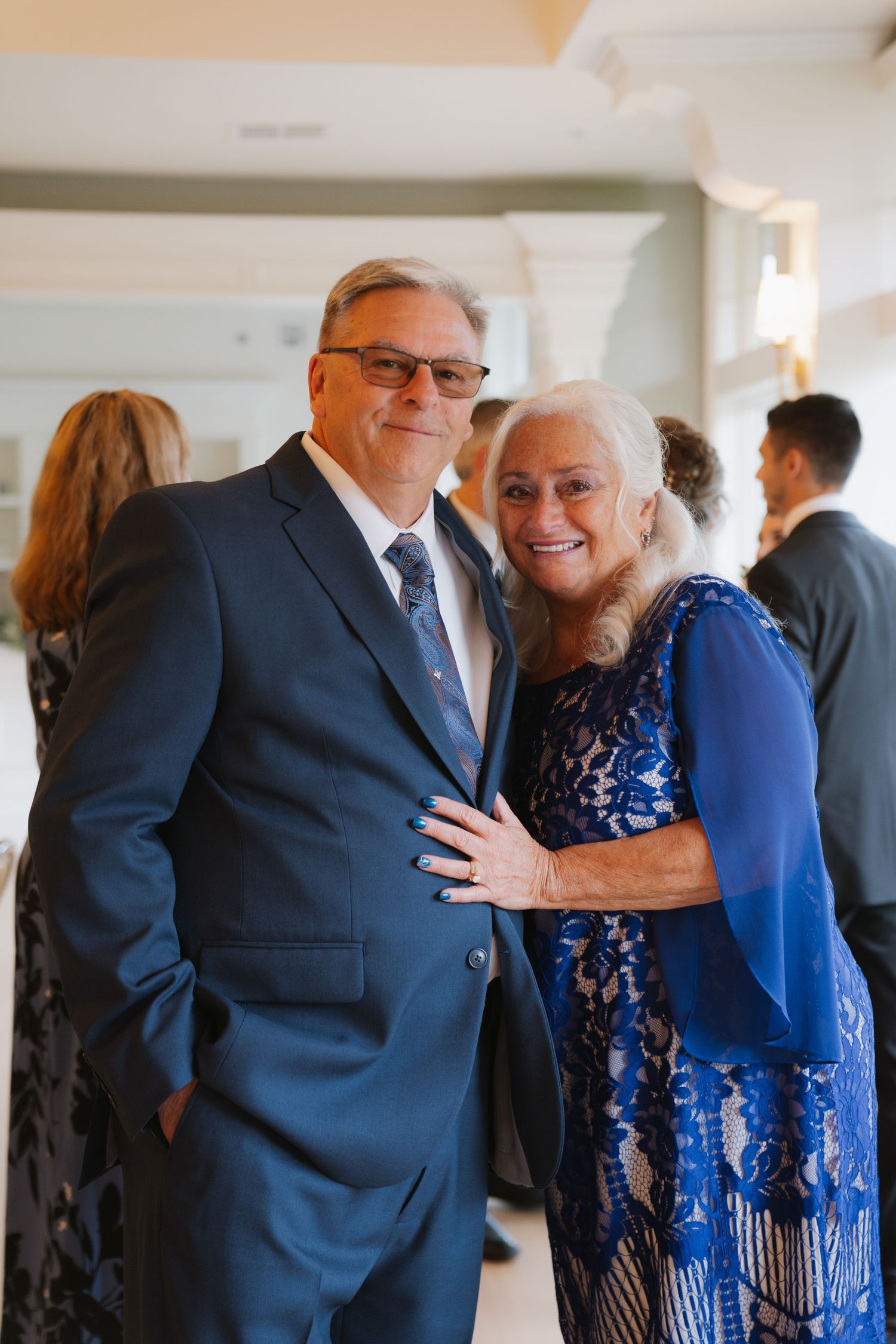 A couple in formal attire share a warm embrace, with the man wearing a navy suit and the woman in a sparkly blue dress.