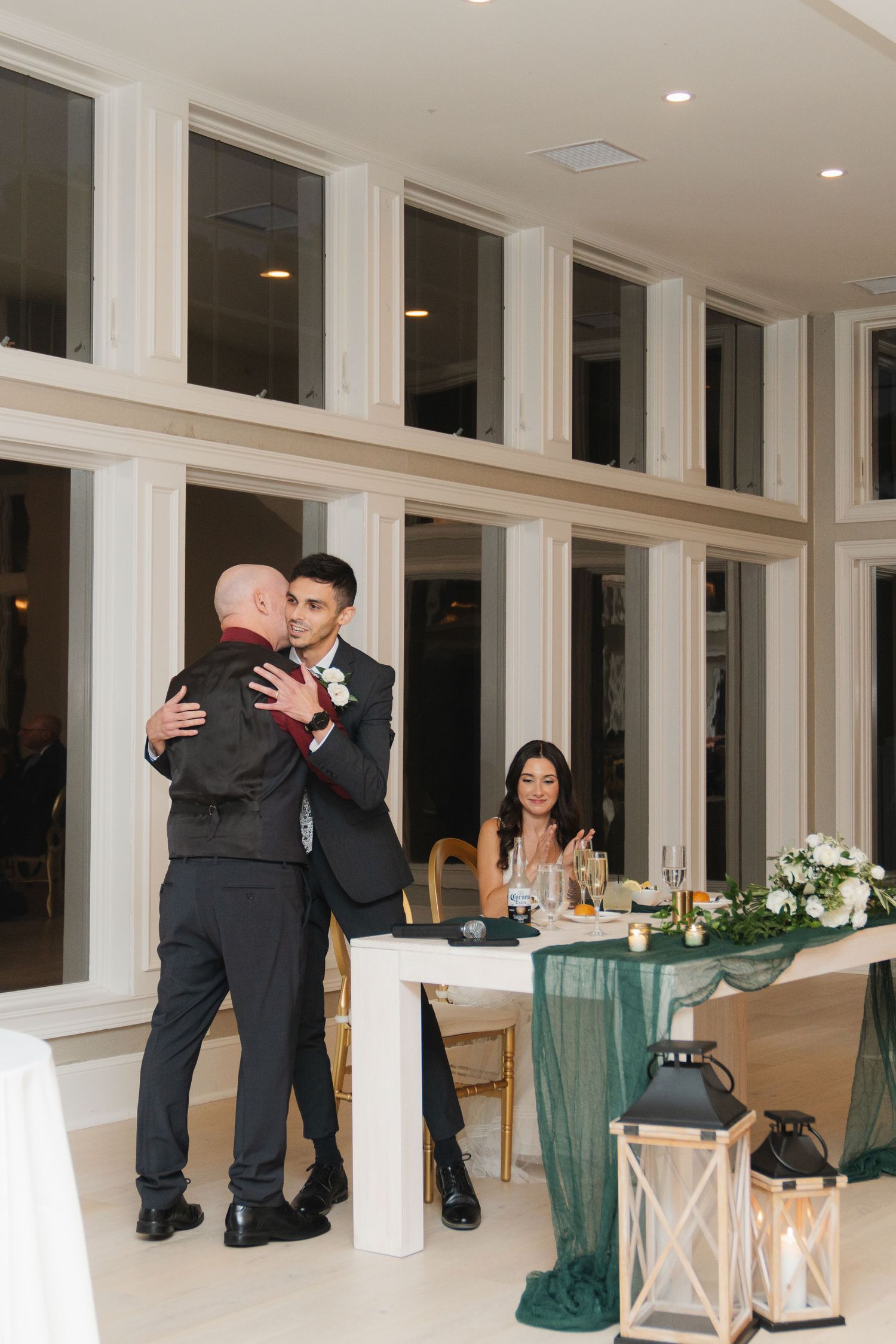 Two people embrace in an elegant reception venue with green tablecloths and white lantern decorations.