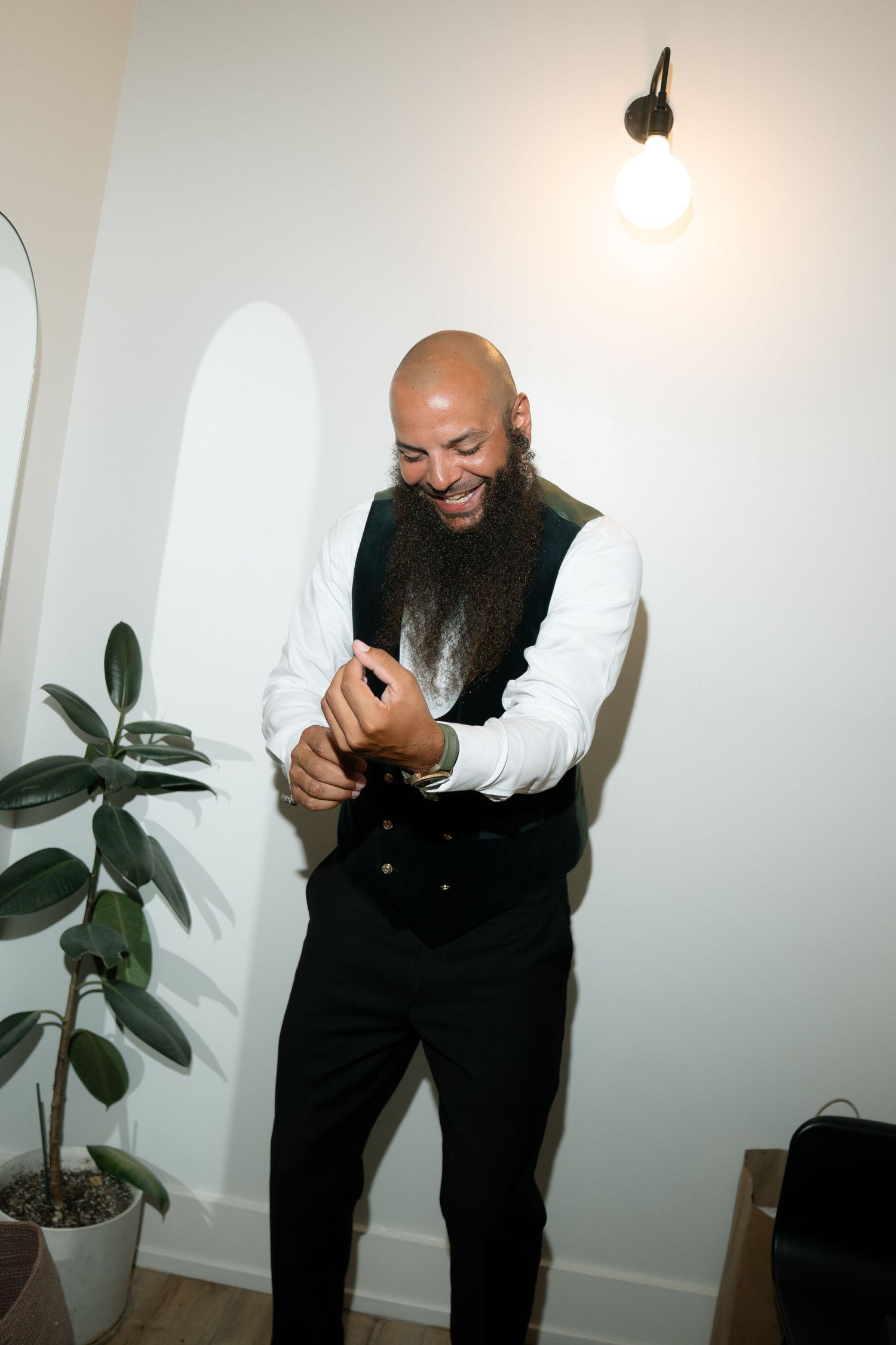 A person in formal attire smiles while adjusting their cuff in a minimalist white room.