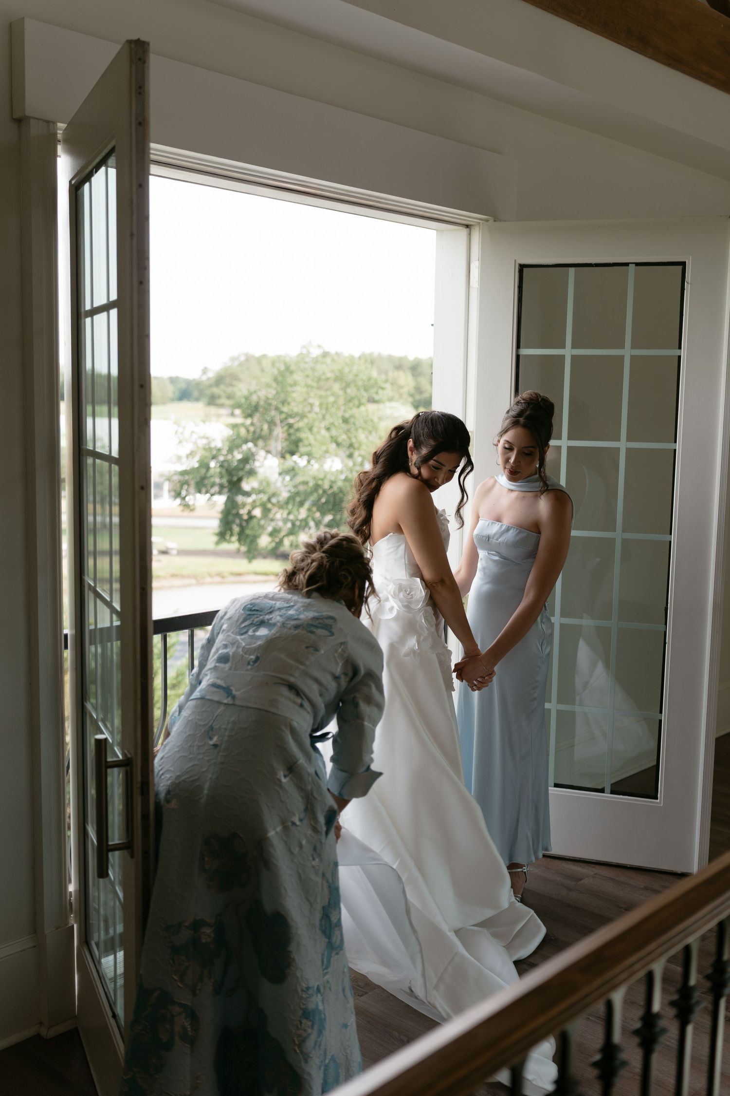 Bride being helped into strapless wedding gown by attendants near French doors on balcony landing.