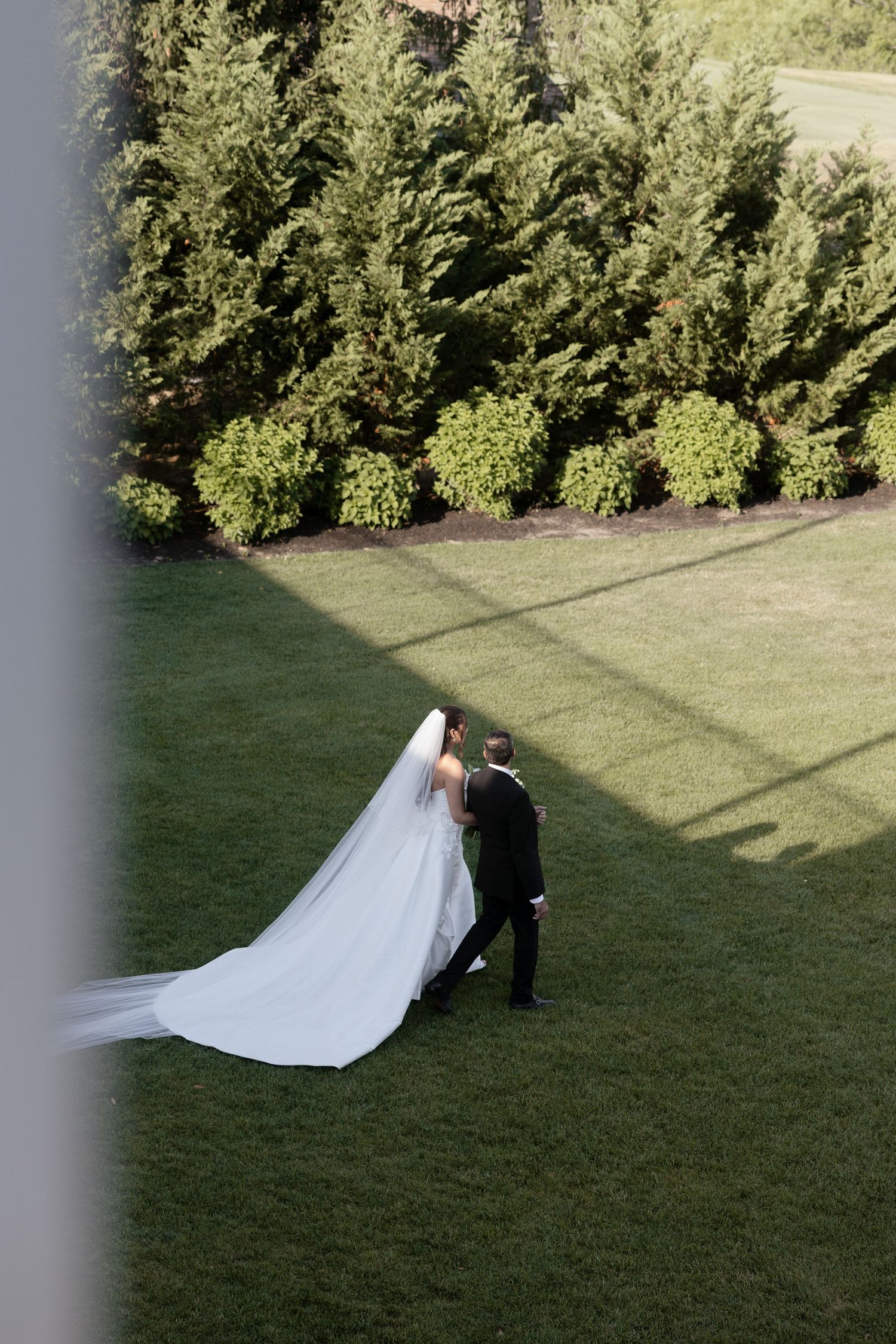 Aerial view of bride and groom walking on green lawn with long flowing veil trailing.