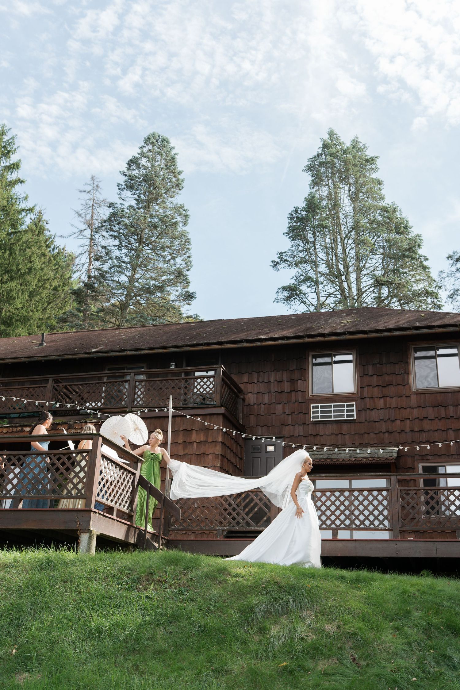 Bride in flowing white gown with long train standing outside rustic wooden lodge surrounded by tall pine trees.