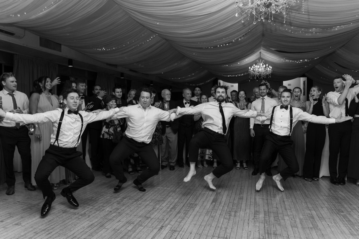 Black and white photo of groomsmen performing a choreographed dance at a wedding reception on a ballroom floor.