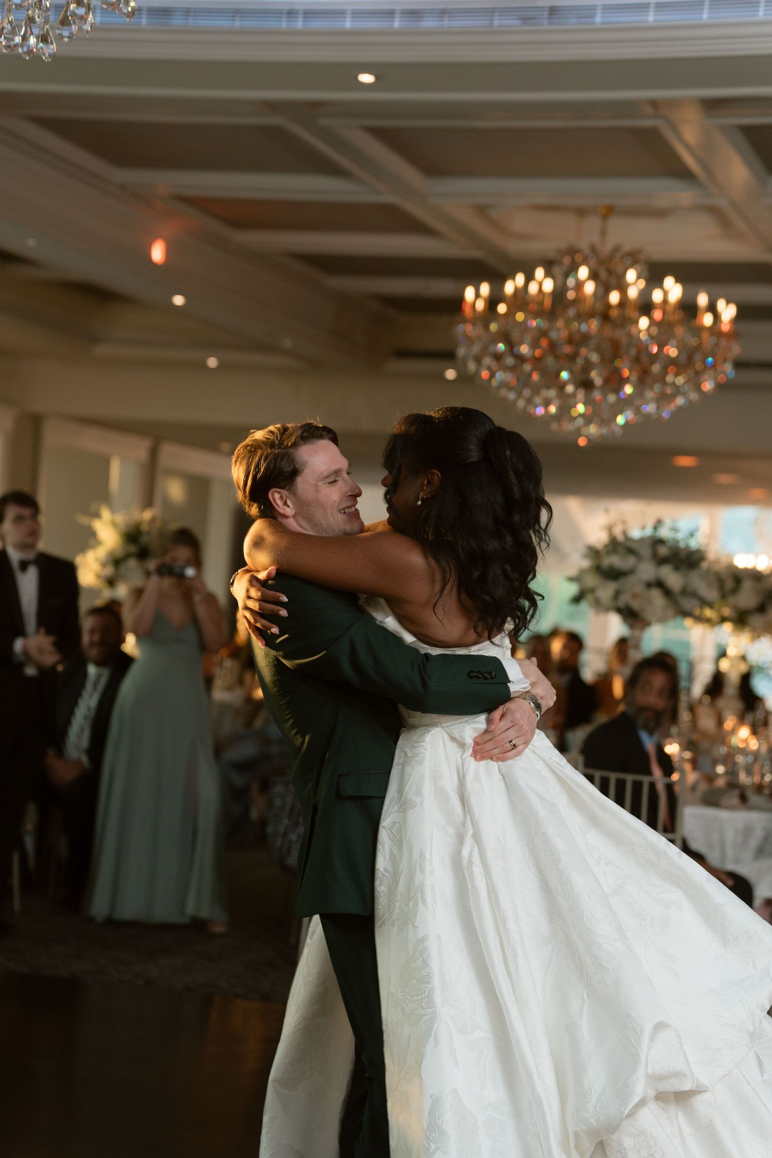 Groom in green suit lifts bride in white gown during first dance at elegant chandelier reception.