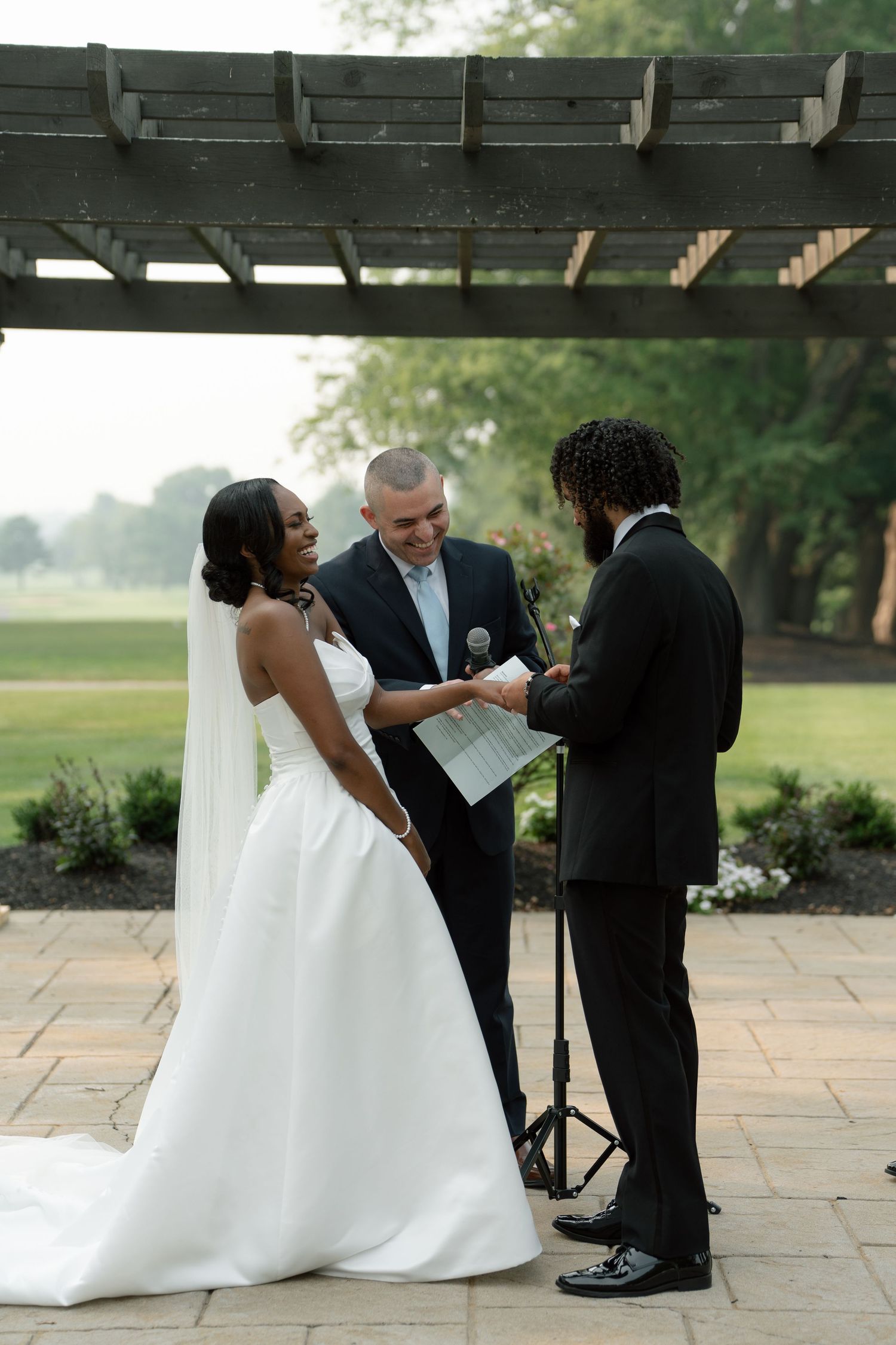 Bride and groom exchange vows under a wooden pergola with an officiant during an outdoor wedding ceremony.