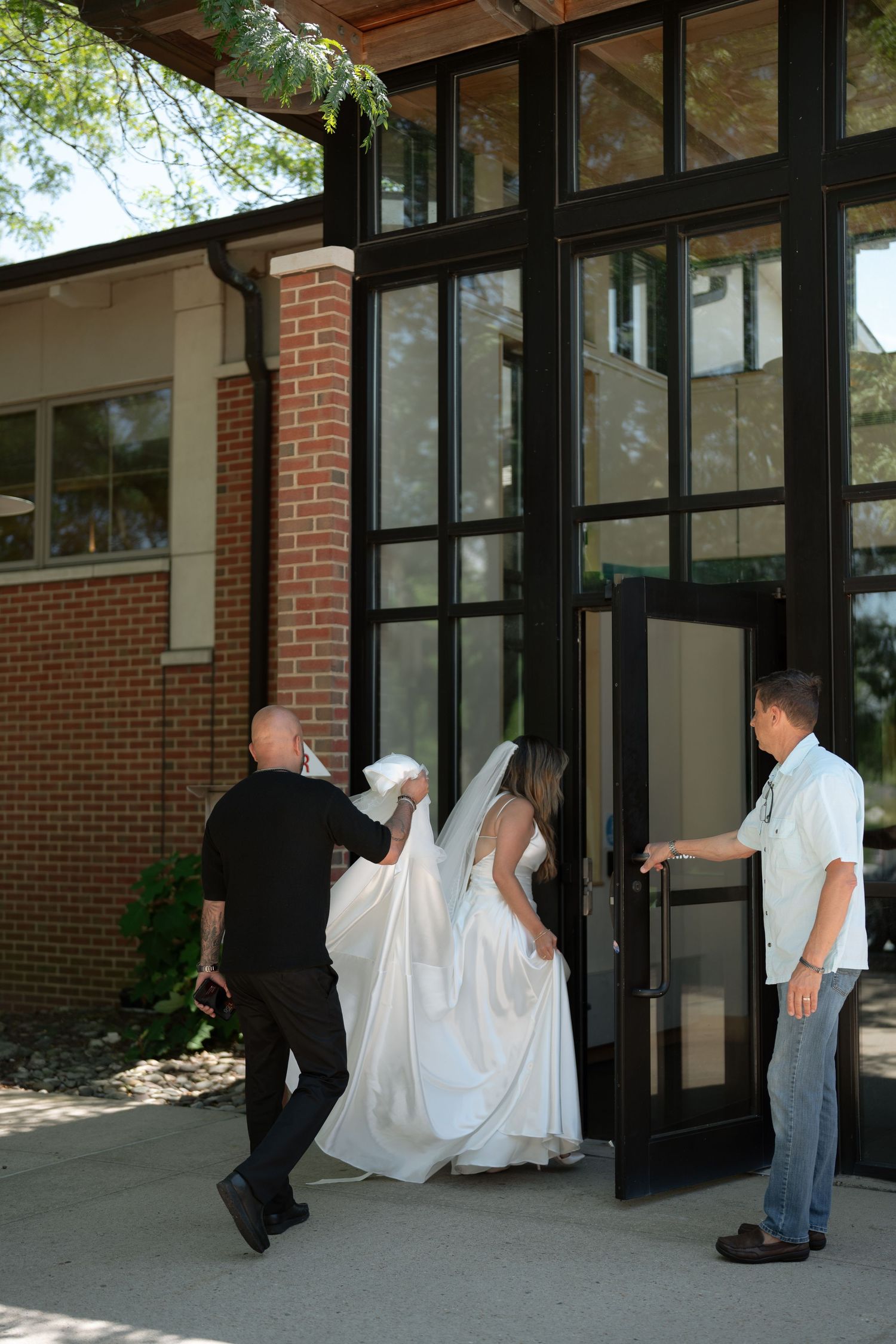 A bride in a white gown and veil enters a brick building assisted by two men holding her dress train.