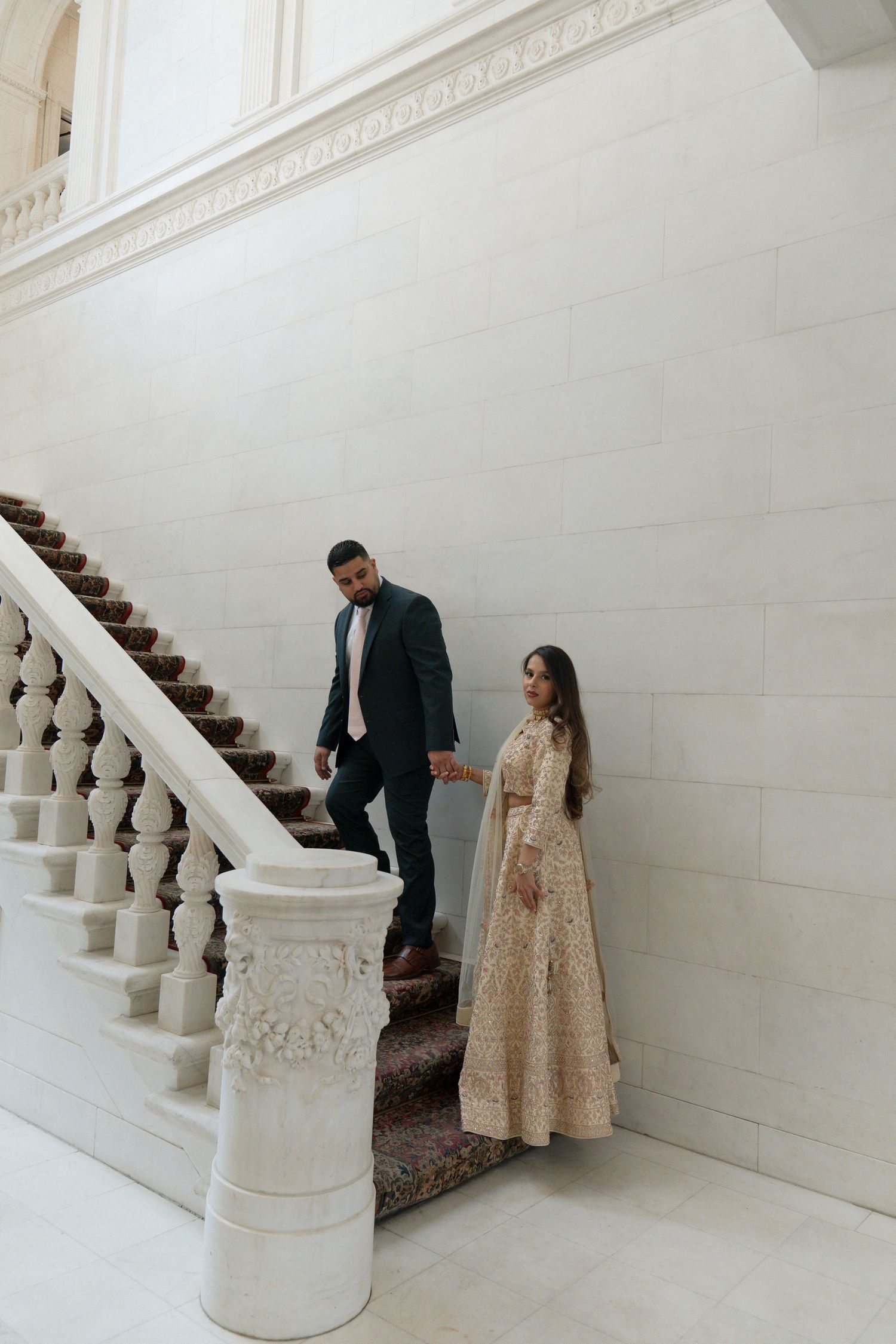 Couple in elegant attire holding hands on ornate white marble staircase, woman in gold embroidered gown, man in dark suit.