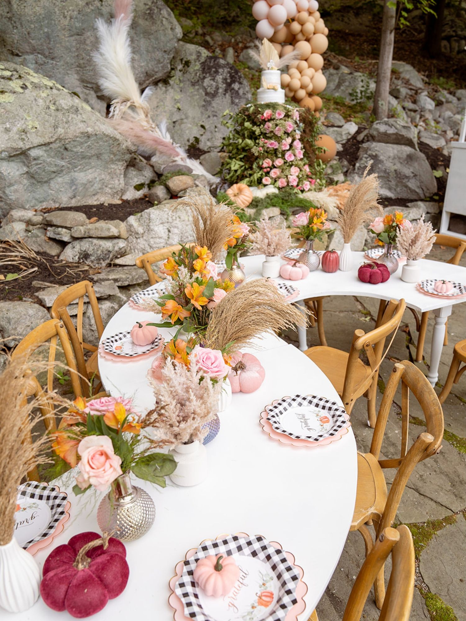 Elegant outdoor table setting with pink and orange florals, velvet pumpkins, and rustic decor against a stone backdrop.