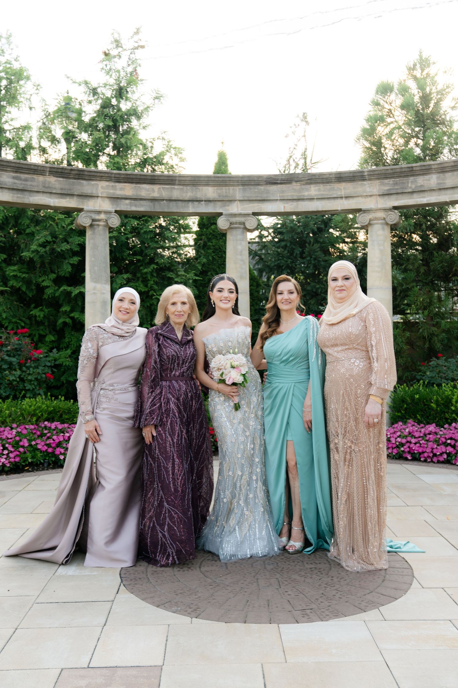 A group poses in formal gowns against a curved stone colonnade with flowers in the background.