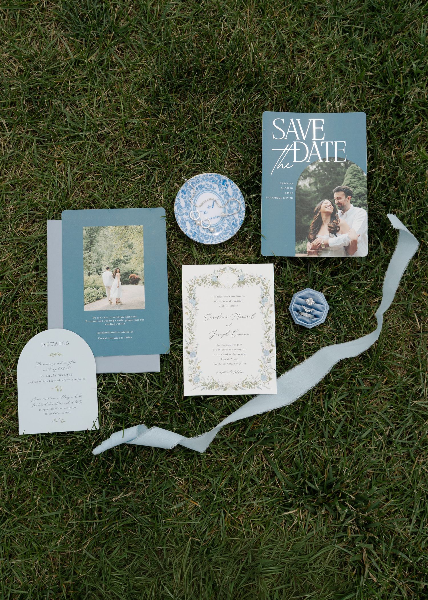 Dusty blue wedding invitation suite with save the date cards, floral details, and ribbon displayed on green grass.