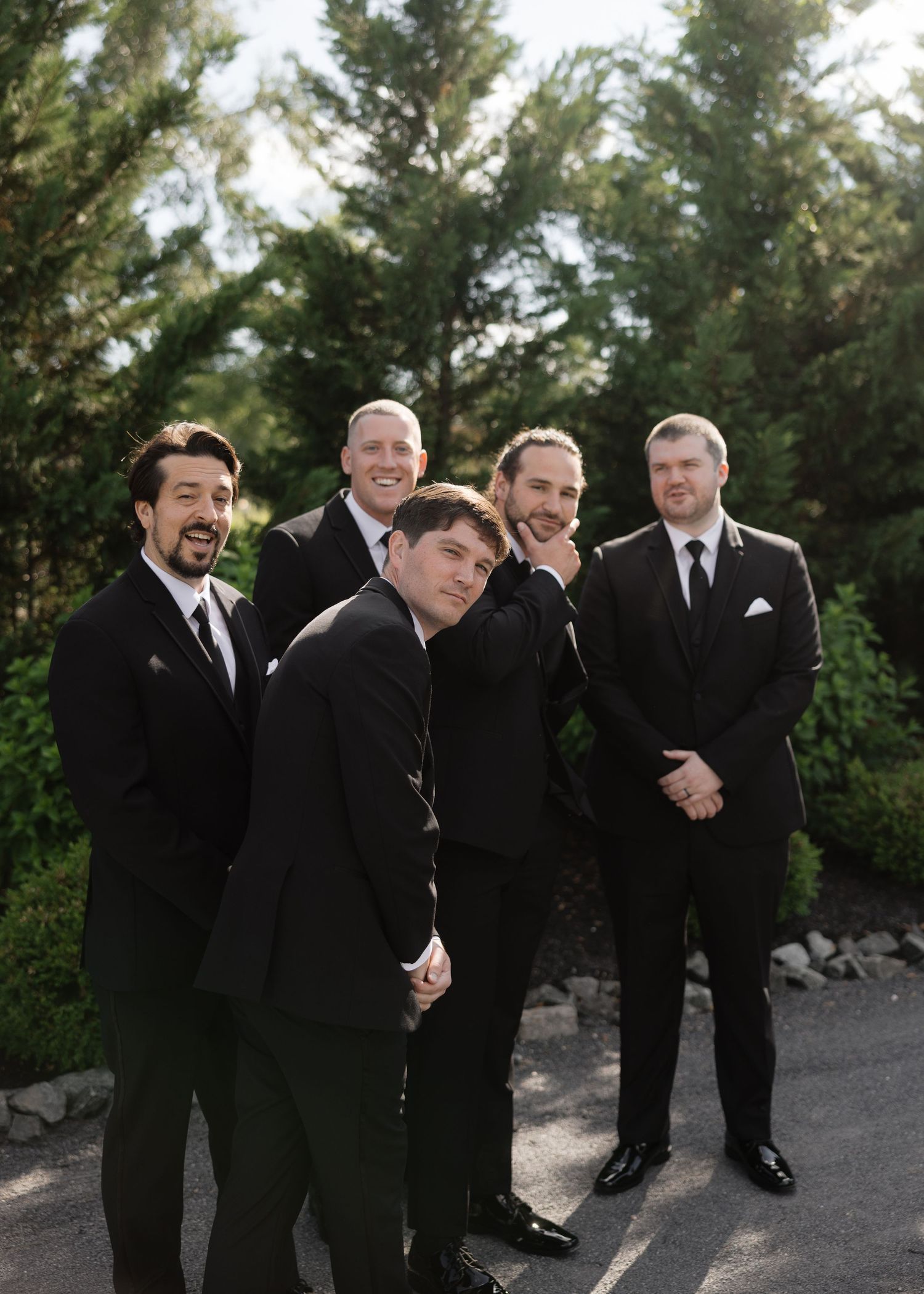Five groomsmen in black tuxedos pose playfully outdoors surrounded by green trees.