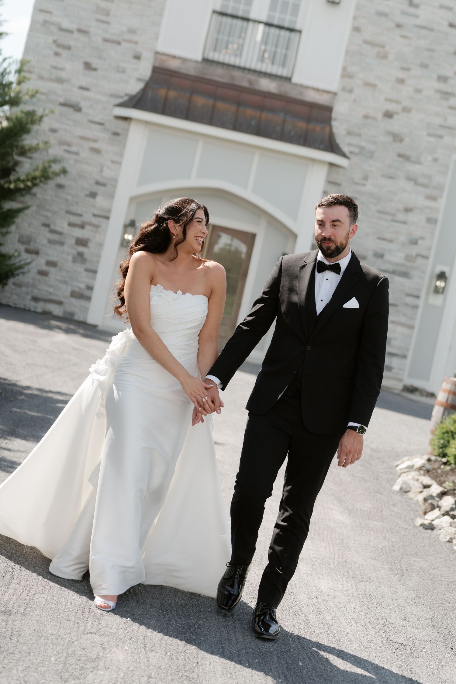 Smiling bride in strapless white gown holding hands with bearded groom in black tuxedo walking outside venue.