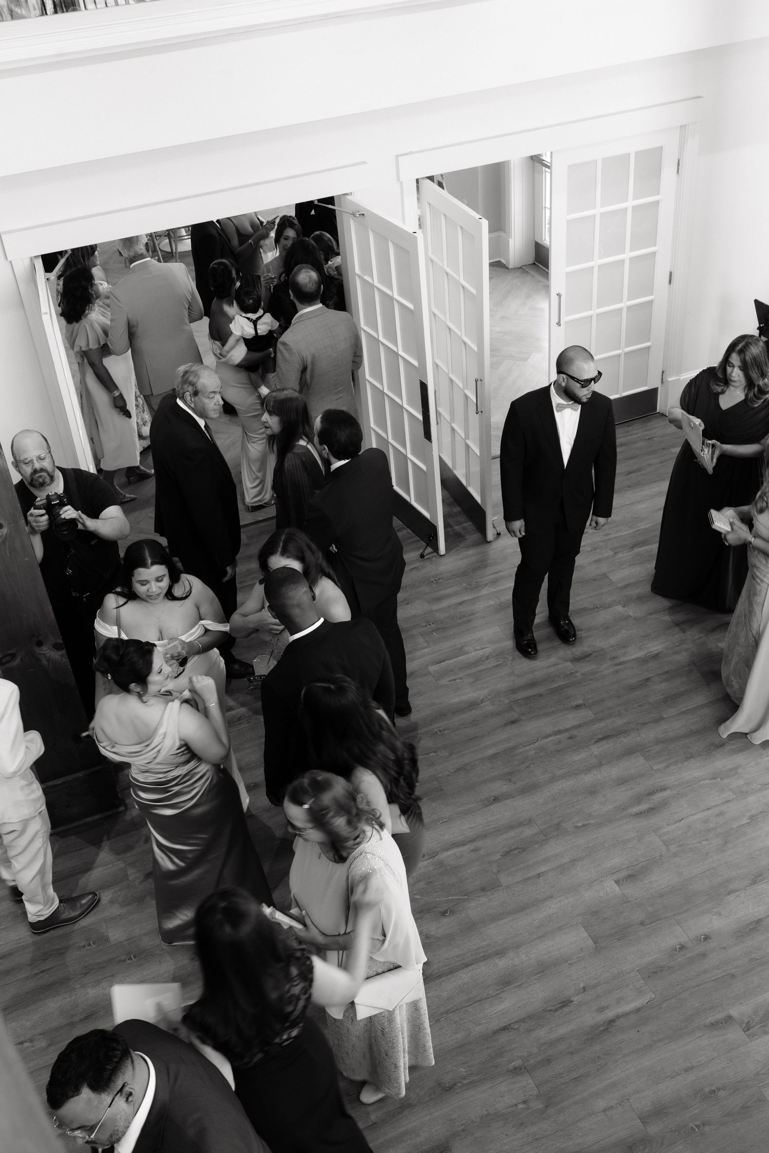 Black and white overhead view of elegantly dressed wedding guests mingling in bright reception hall.