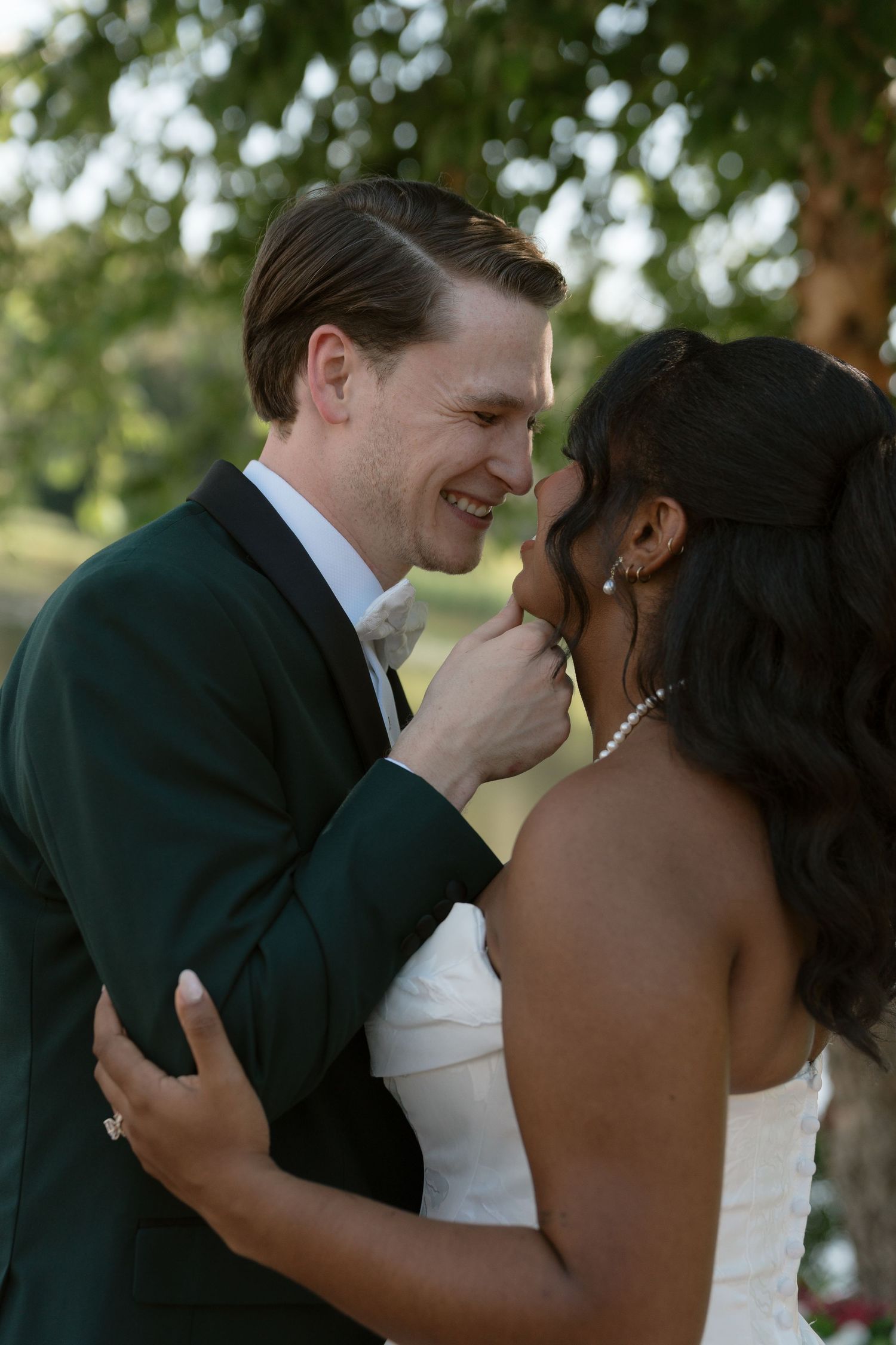Interracial couple sharing an intimate moment on their wedding day, groom in green tuxedo smiling at bride in white gown.