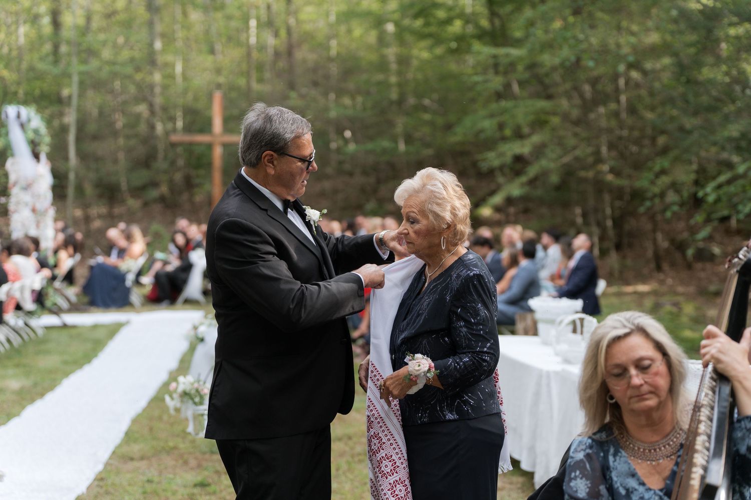 Outdoor wedding ceremony with guests seated, officiant in tuxedo assisting elderly woman near floral arch in wooded setting.
