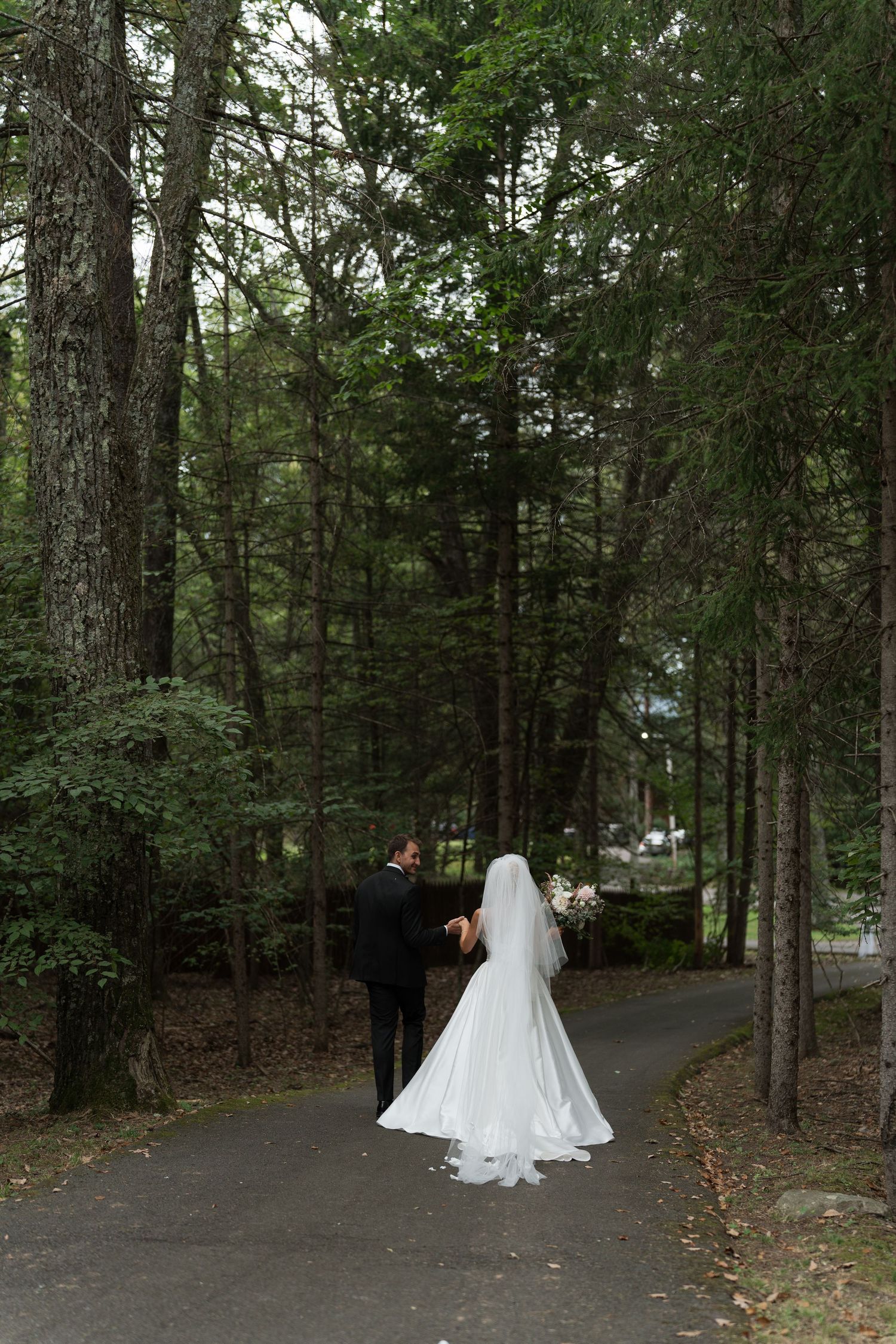 Bride and groom walking hand in hand along a tree-lined path in a lush green forest setting.