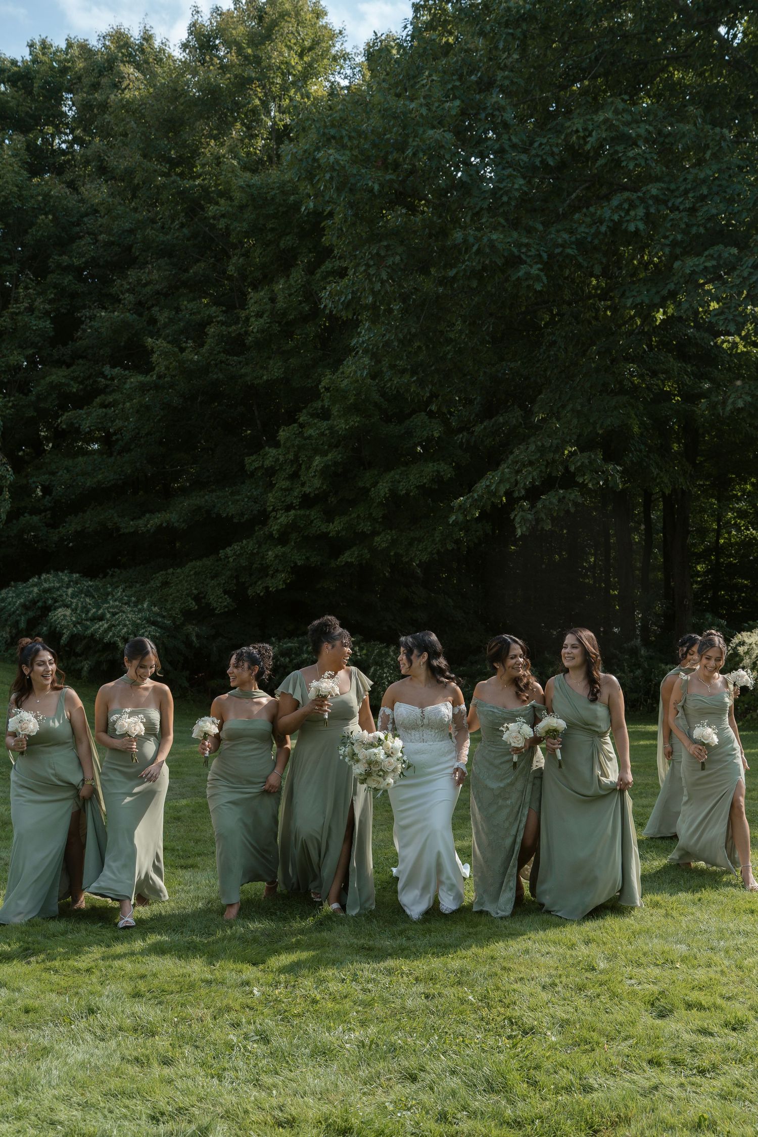 Bride in white gown walks with bridesmaids in sage green dresses carrying white bouquets on green lawn.